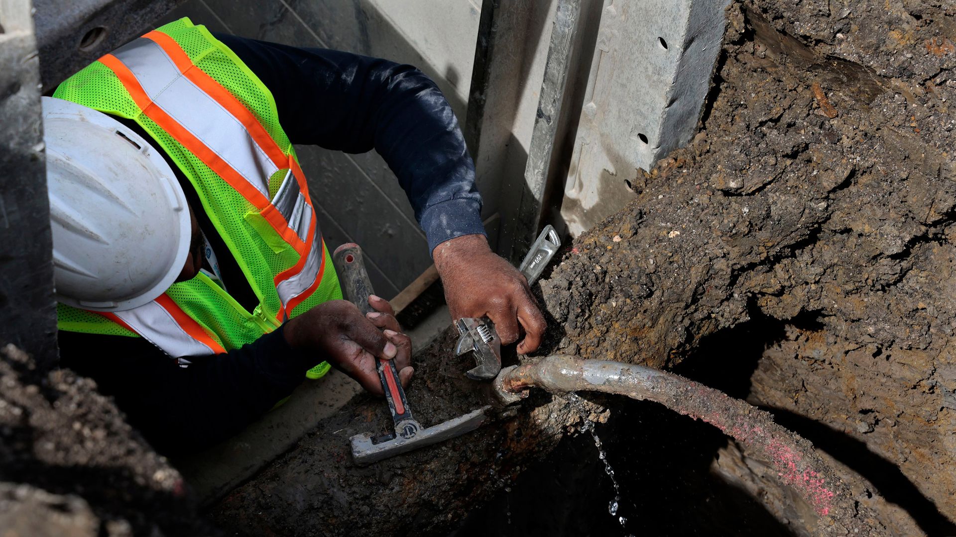 A person in a yellow construction vest and white helmet uses a tool on a pipe in a hole surrounded by dirt