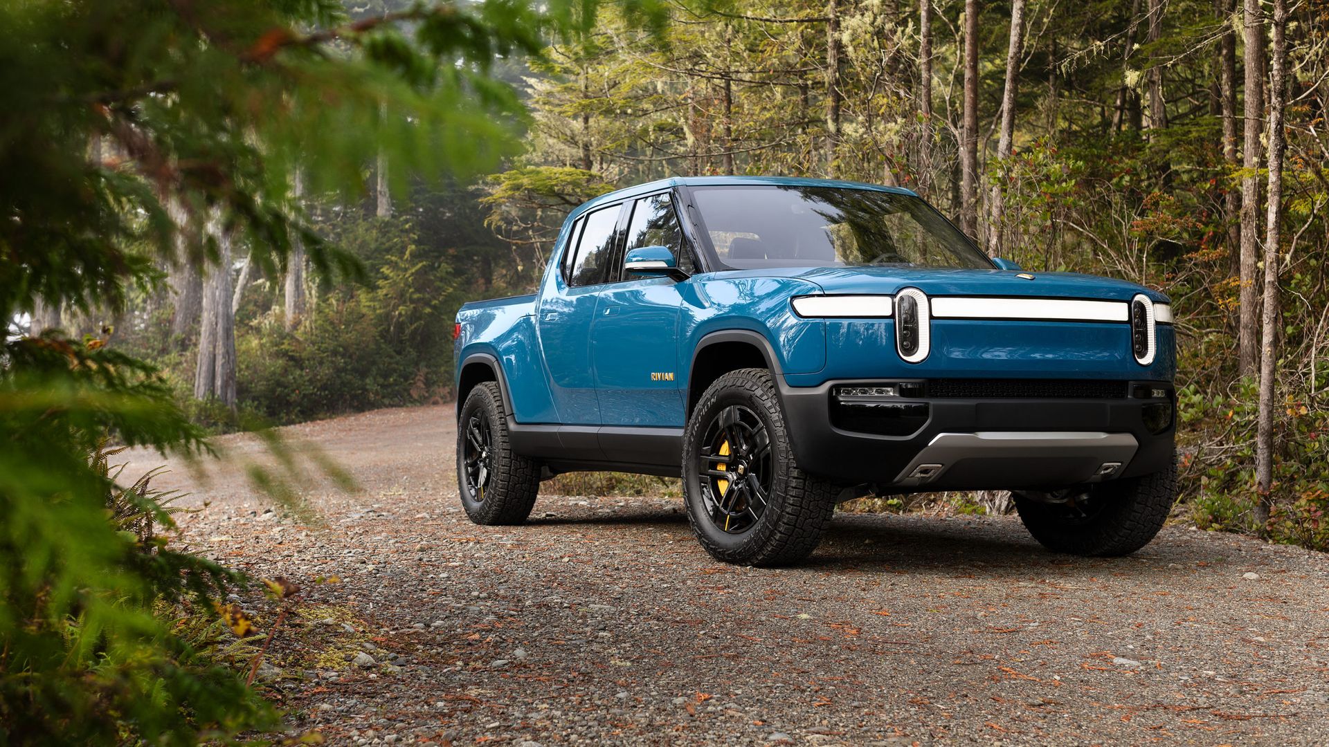Image of a blue Rivian R1T electric pickup truck on a gravel road in the woods. 