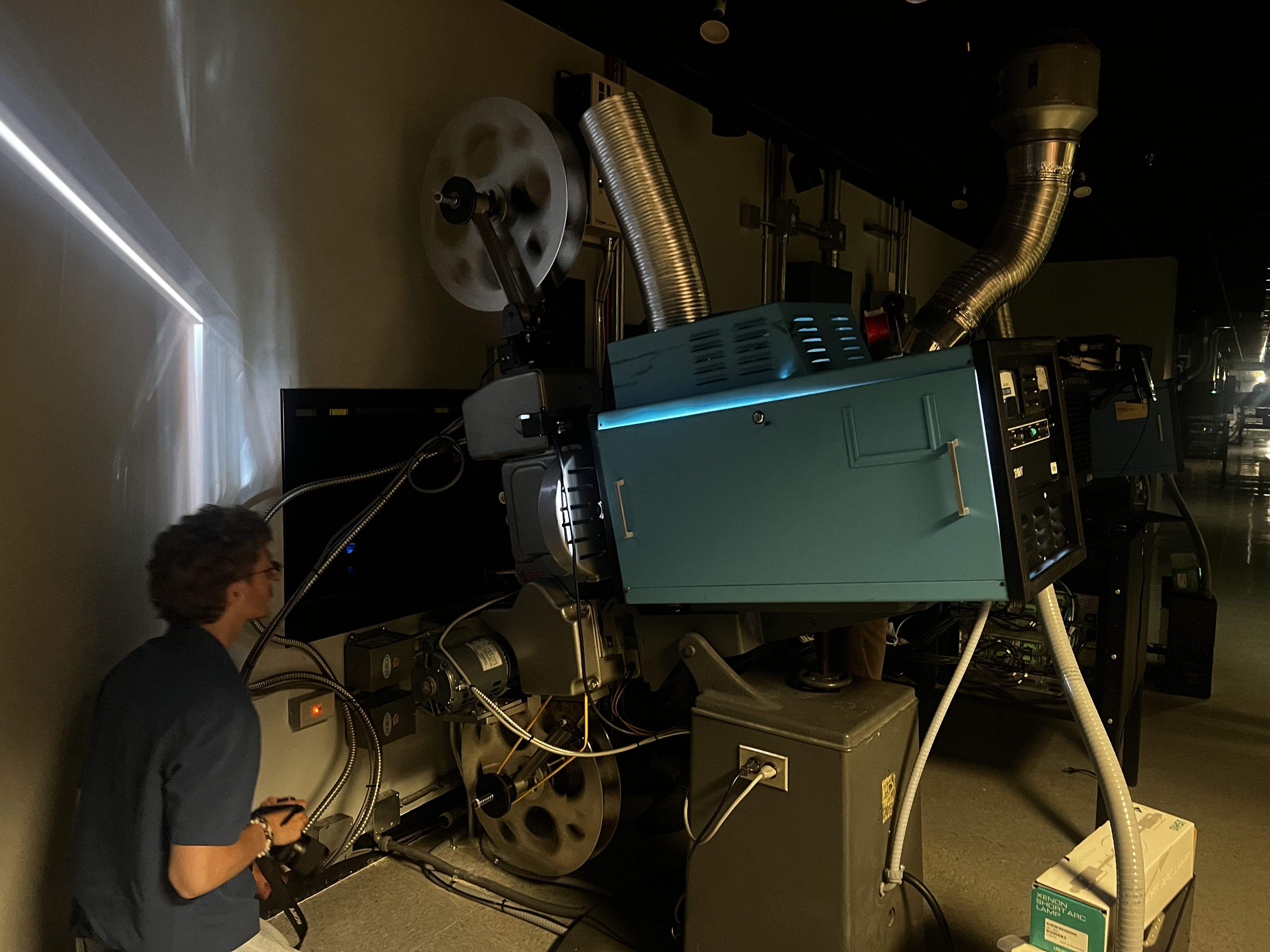 Dim lab with teal industrial equipment and large silver ducts. A person in a blue shirt stands beside tangled cables near a control panel and a circular metal wheel.