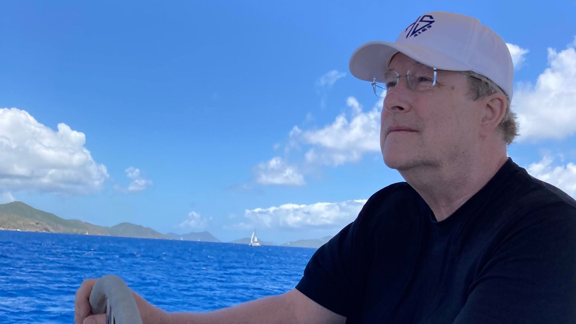 A man wearing a white Adidas cap and glasses sits at a boat railing, gazing toward the horizon over a bright blue sea with distant mountains and a small sailboat in the background.