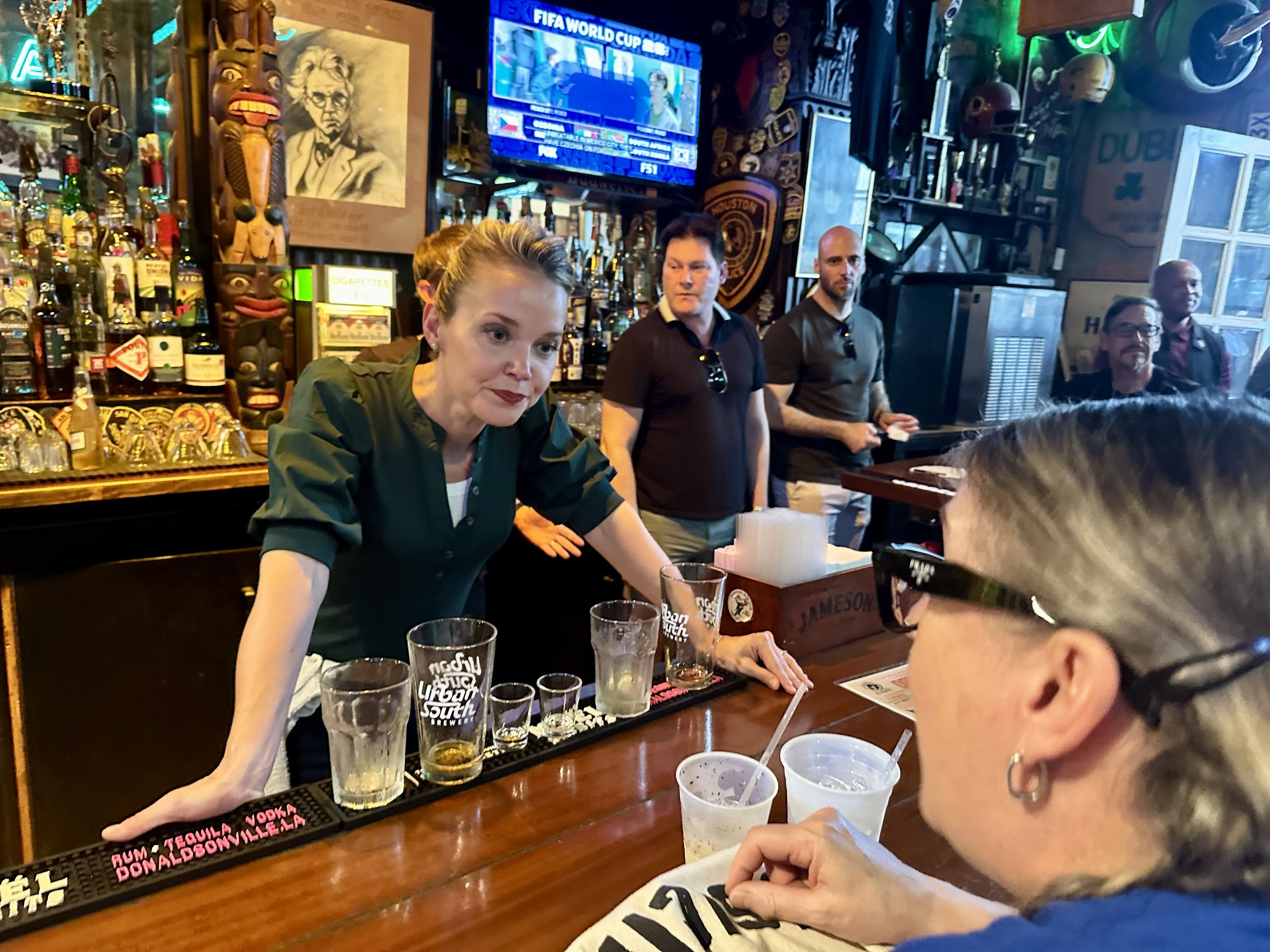 Bartender in a green shirt leans over a busy bar, with rows of glassware and liquor bottles behind her, a carved tiki figure, neon signs, and a TV showing a sports broadcast in the background.