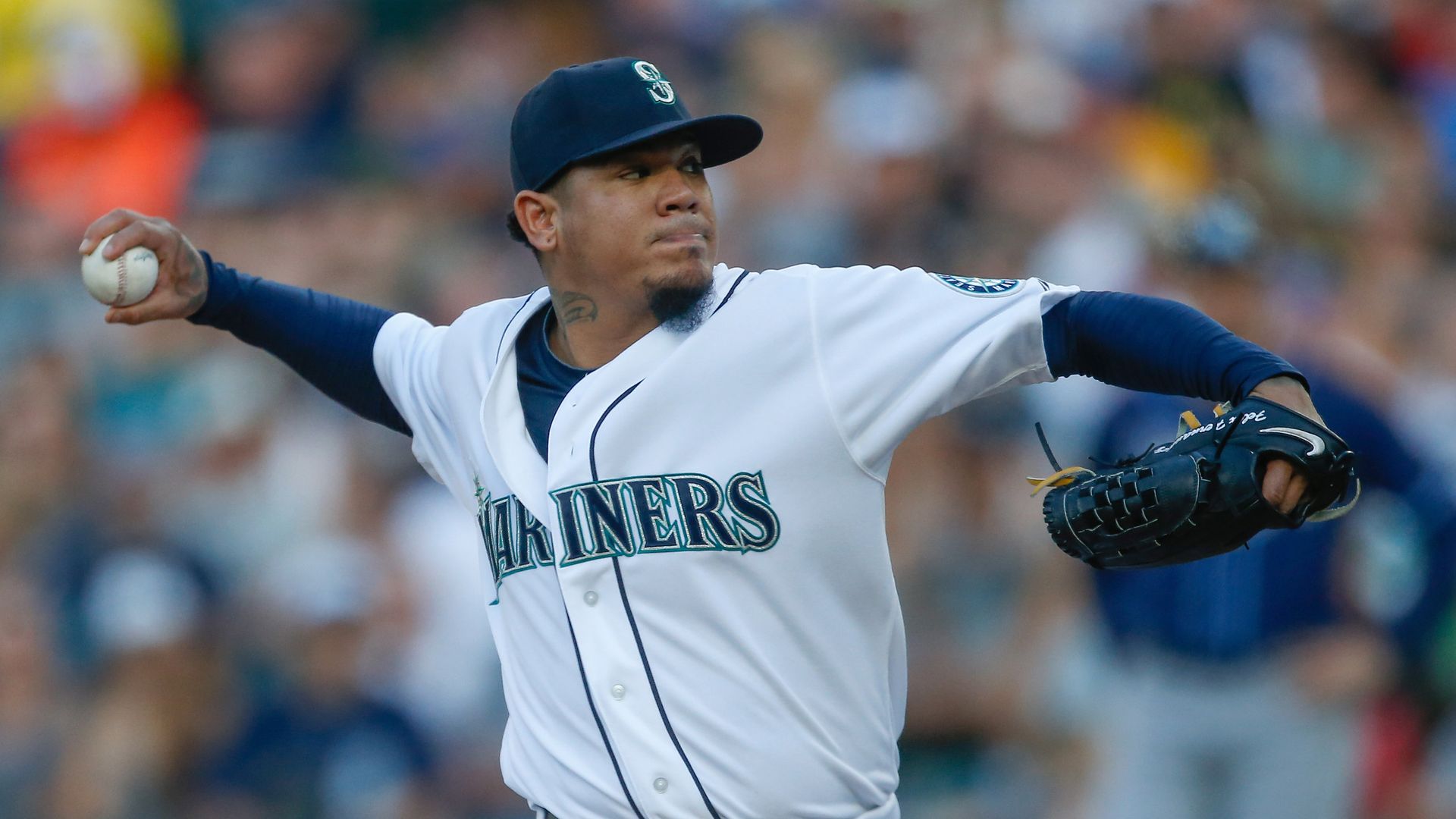 A man in a Mariners jersey and cap pulls back to wind up before throwing a pitch. 