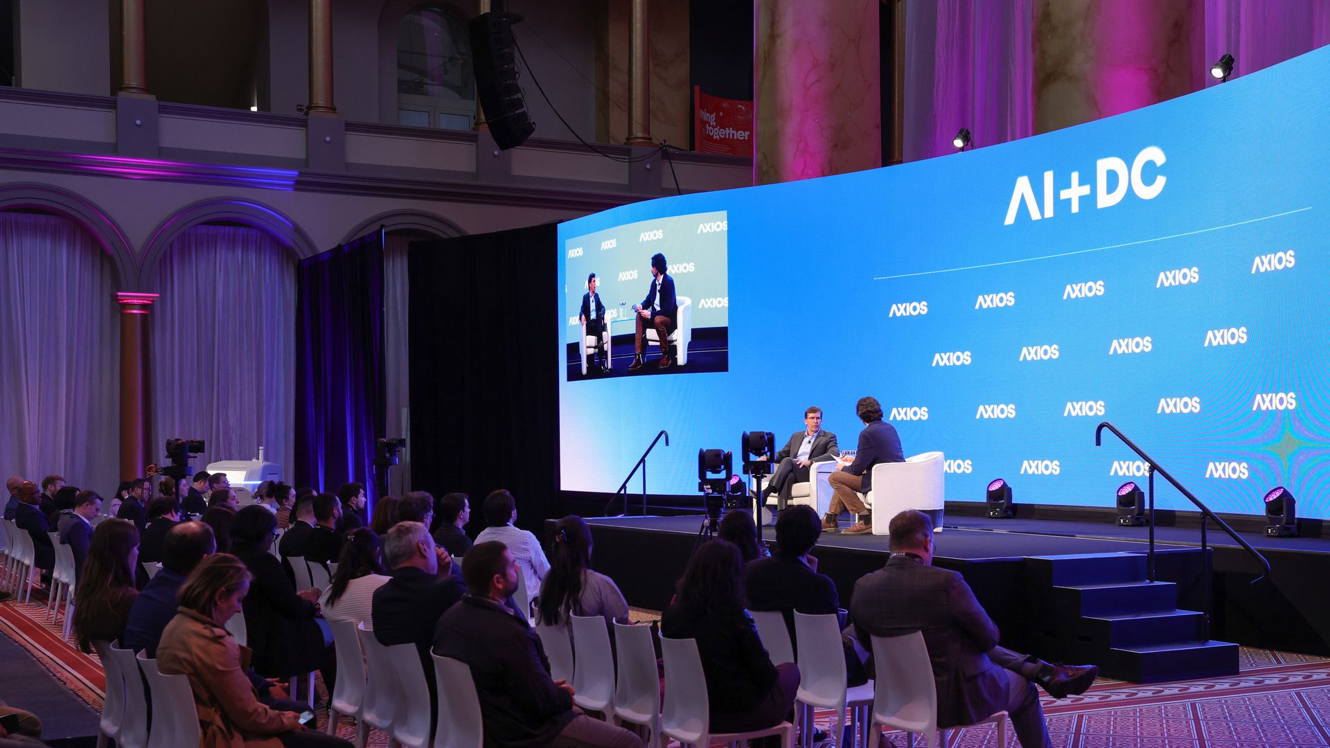 A conference stage with two men seated in white chairs, speaking into microphones. A large blue screen reads "AI+DC" with repeated "AXIOS" logos; an audience watches in a grand, purple-lit hall.