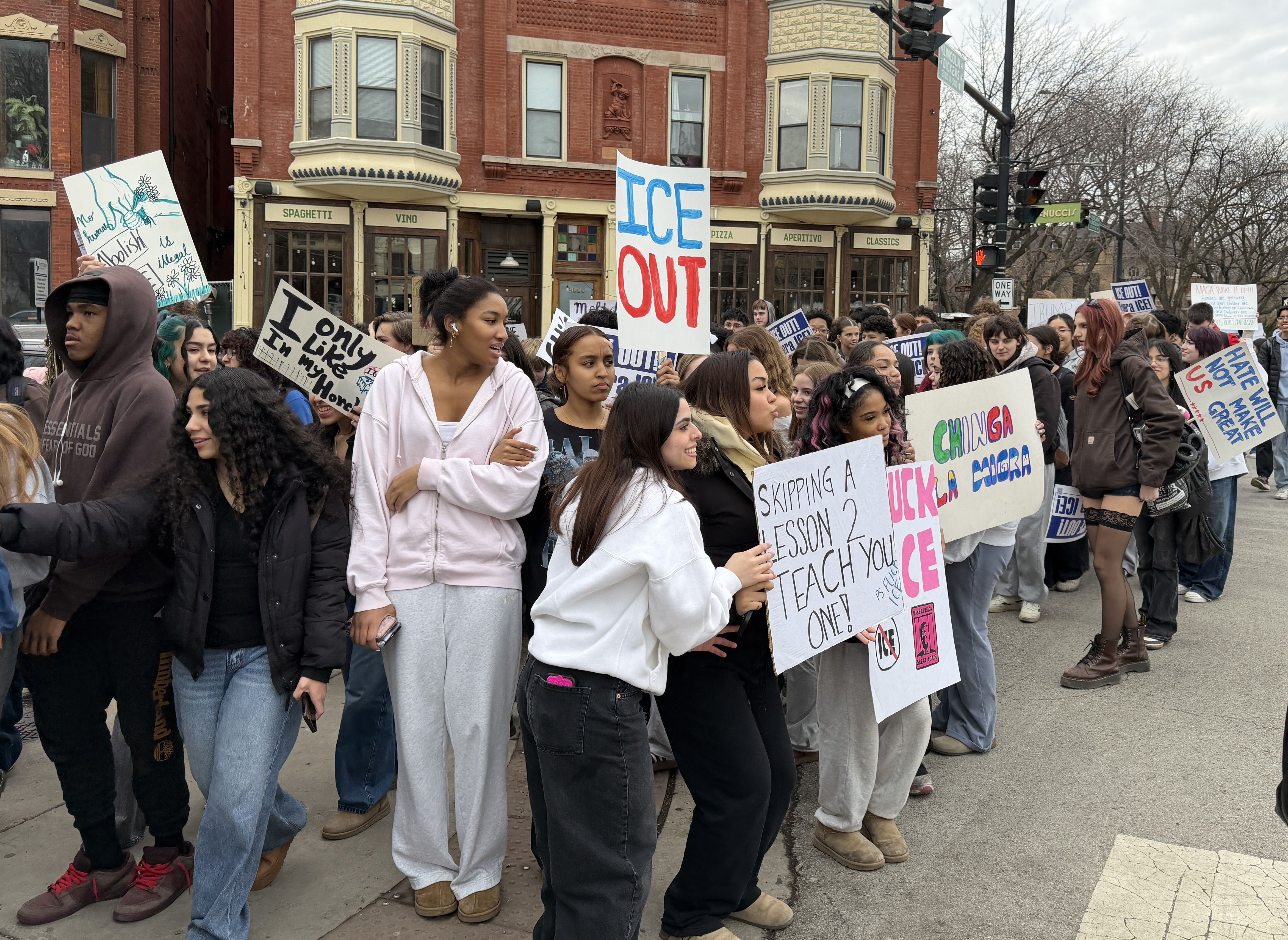 Crowd of young protesters holding signs with messages like "ICE OUT" and "Skipping a lesson 2 teach you one" outside a brick building with bay windows on a cloudy day.