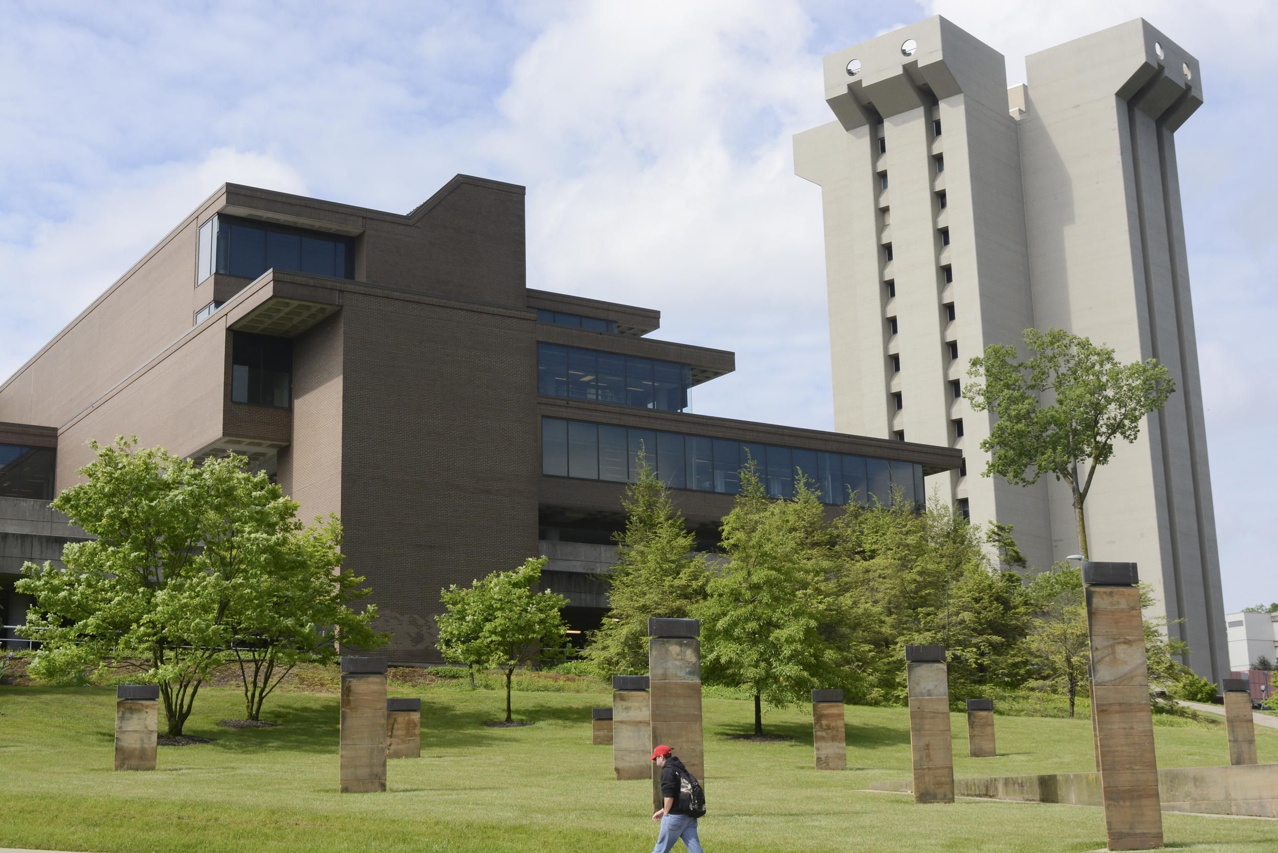 University of Cincinnati campus scene with a brown brick building on the left and Crosley Tower - a tall gray concrete structure - on the right, blue sky, trees, and a person in a red cap walking on the lawn.