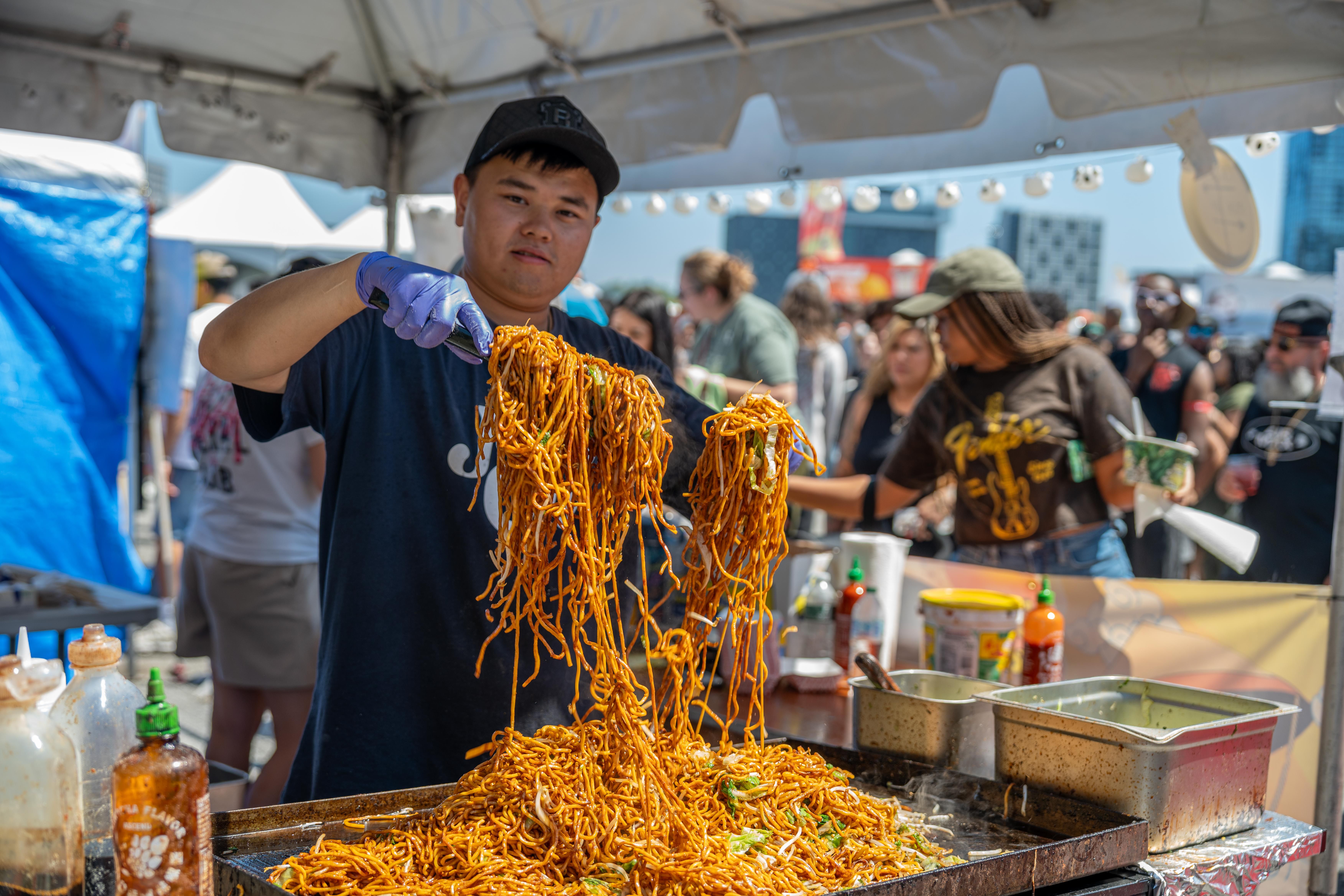 A street food vendor in a black cap and blue gloves lifts a tangle of saucy noodles from a grill, outdoors under a canopy as a crowd of festival-goers watches.