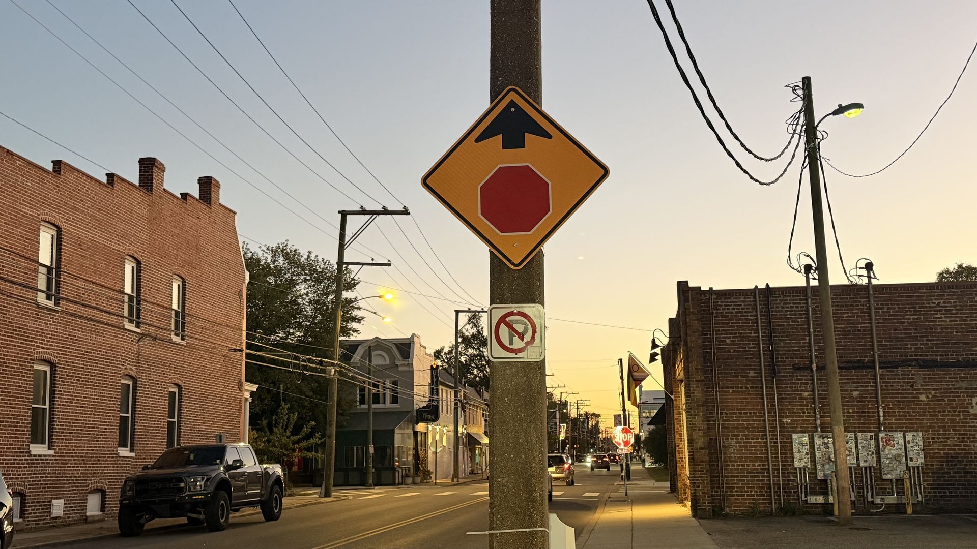 Yellow diamond traffic sign with a black upward arrow and a red octagon inside, mounted on a concrete pole in a brick-block street at sunset, with cars and buildings nearby.