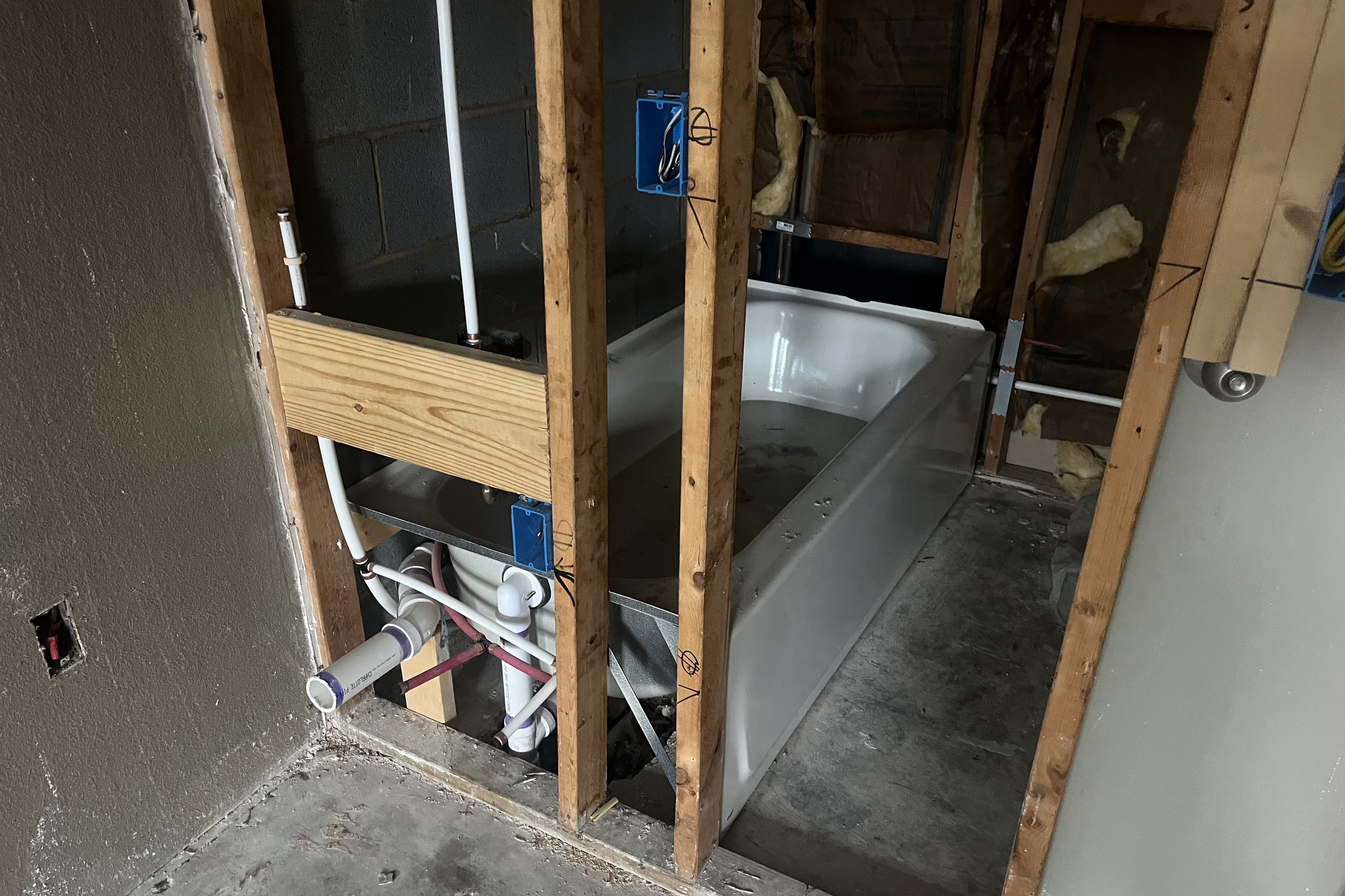 Bathroom under renovation showing exposed wooden studs, plumbing pipes, electrical boxes, and a white bathtub installed but surrounded by unfinished walls and concrete floor.