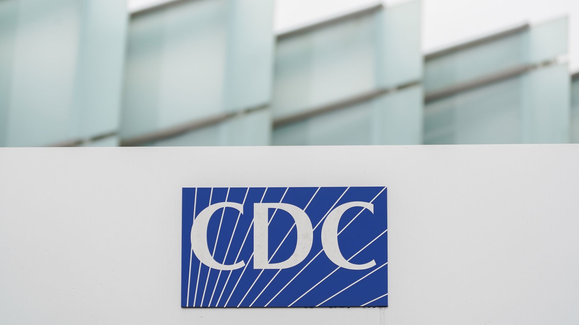 Blurry exterior of a building with tall pale blue glass panels rising above a white wall, and a blue square sign with white diagonal lines and large white lettering visible near the bottom that read, CDC.