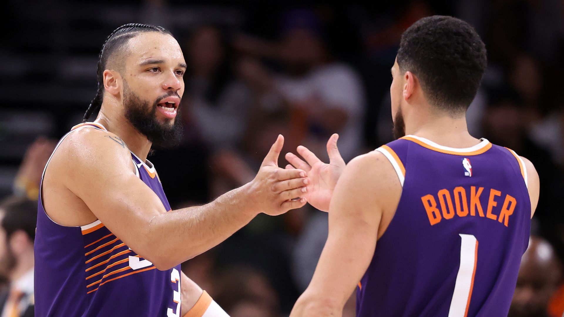 Two basketball players in purple jerseys, one with his back to the camera and the word "Booker" on the back," slap hands on the court, with blurry images of people in the seats behind them. 
