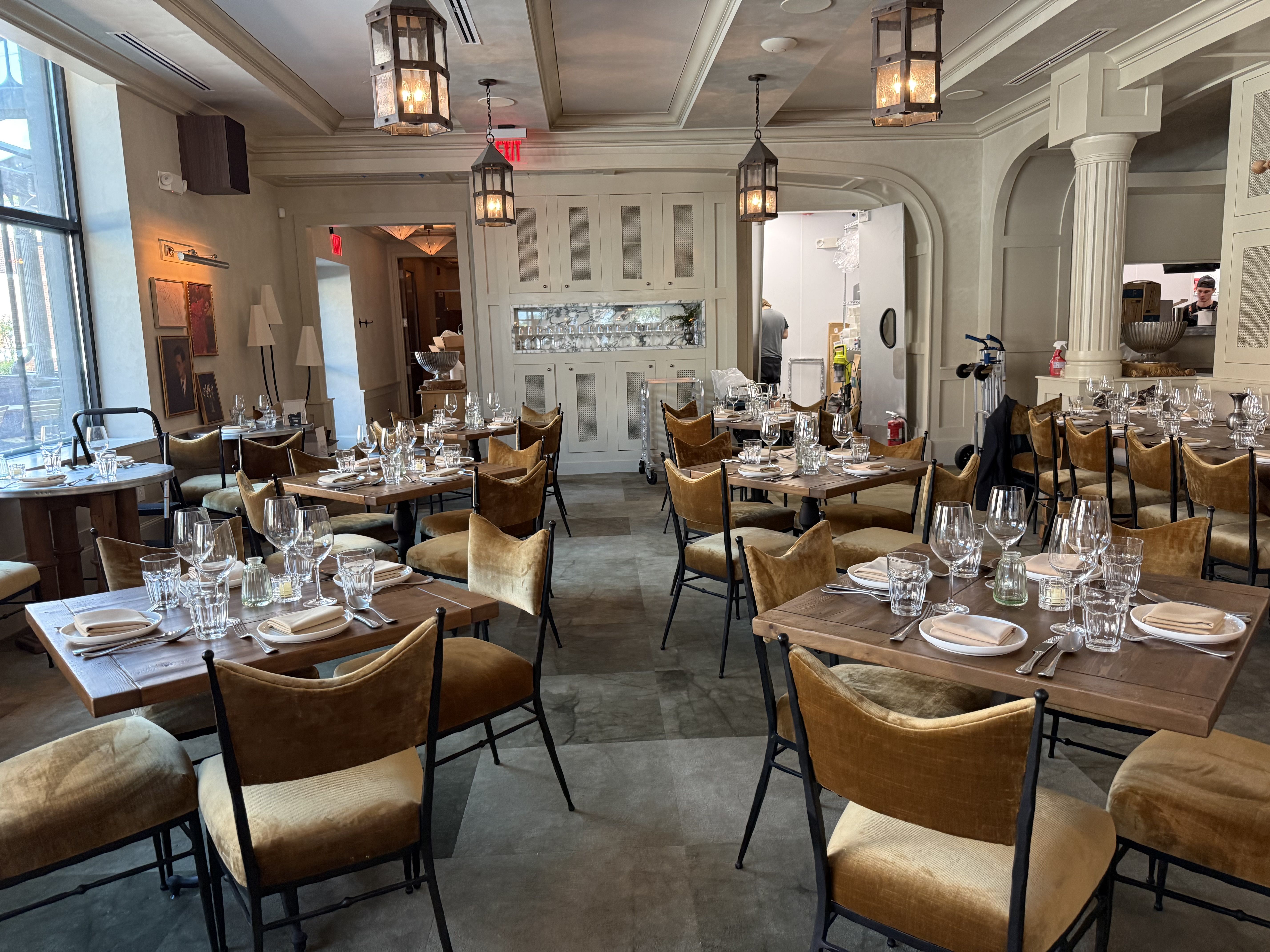 Spacious, softly lit restaurant dining area with wooden tables set with glassware and plates, tan chairs, hanging lantern lights, large windows, and an open kitchen in the back.