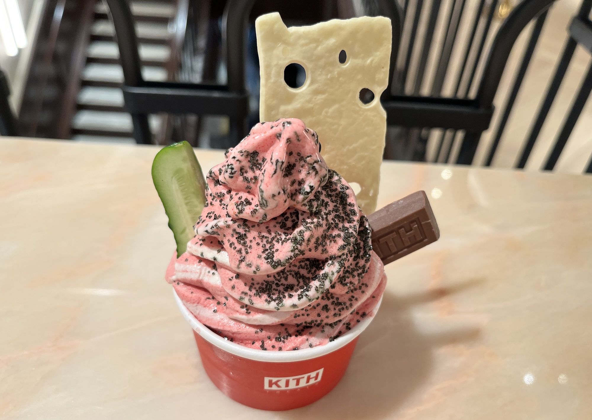 Red KITH cup holds pink swirled soft-serve with black sprinkles, topped by a beige cheese-shaped wafer and a cucumber slice, plus a chocolate bar; light tabletop and striped chairs in the background.