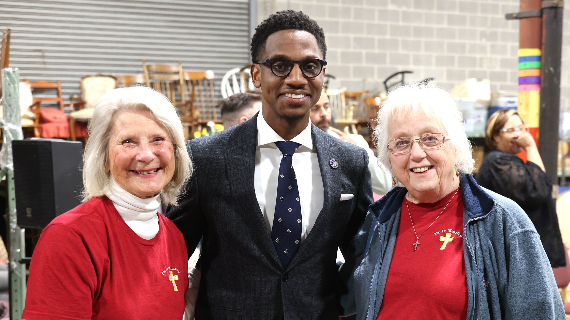 Black bespectacled man in suit stands between two elderly white women in red t shirts.