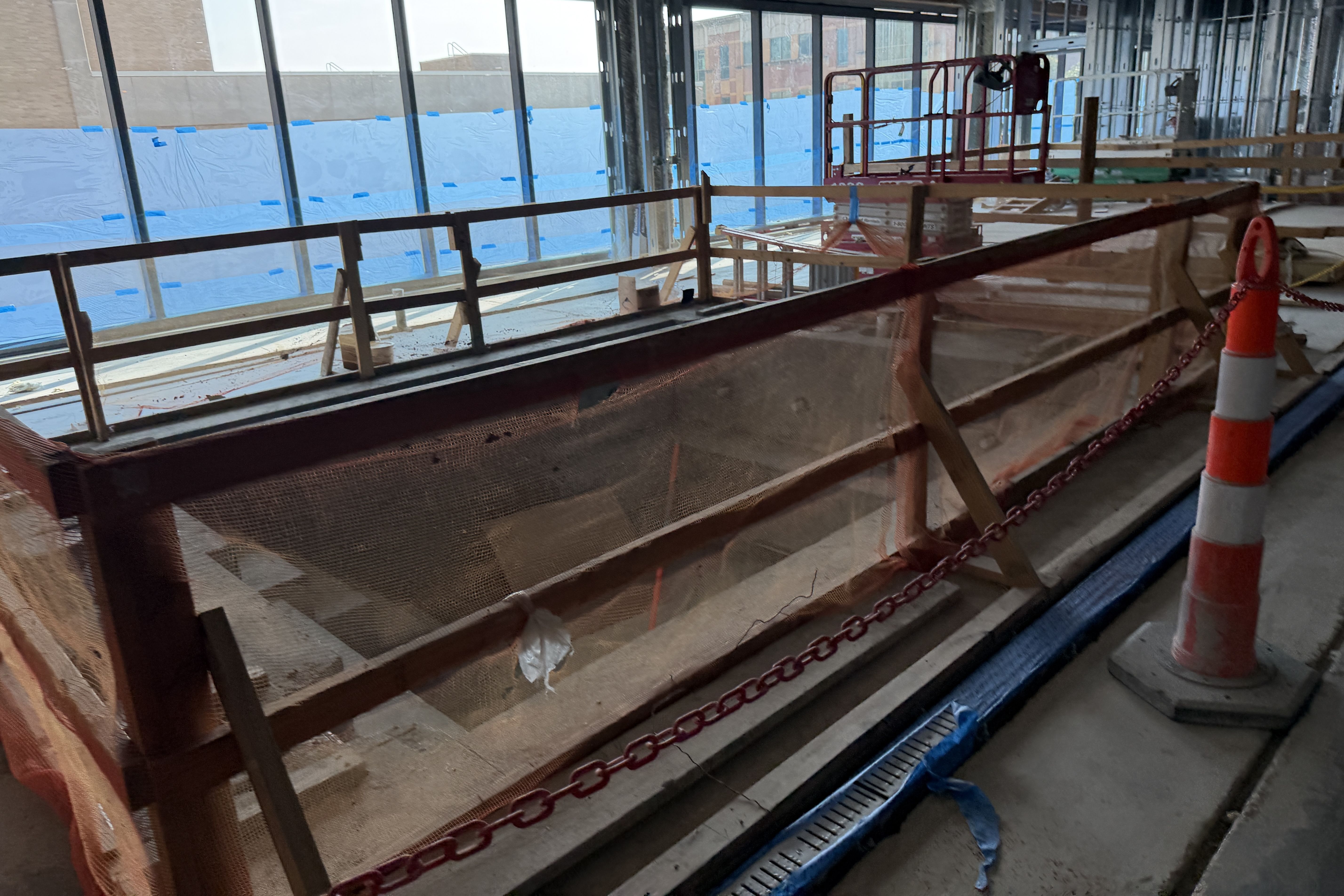 Interior construction site with exposed framework, wooden guardrails, scaffolding, and blue protective plastic along large glass windows; orange safety cone and red chains mark a restricted area.