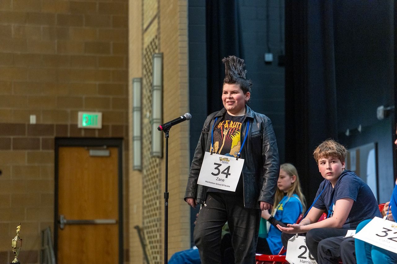 Person with a tall black mohawk, black leather jacket, and bib number 34 stands at a microphone on a stage; seated peers watch in a brick-walled venue.