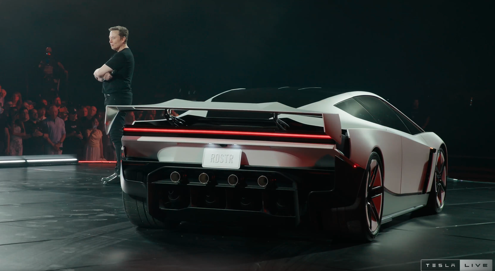 Elon Musk stands with his arms crossed beside the new Tesla Roadster sports car on a stage.