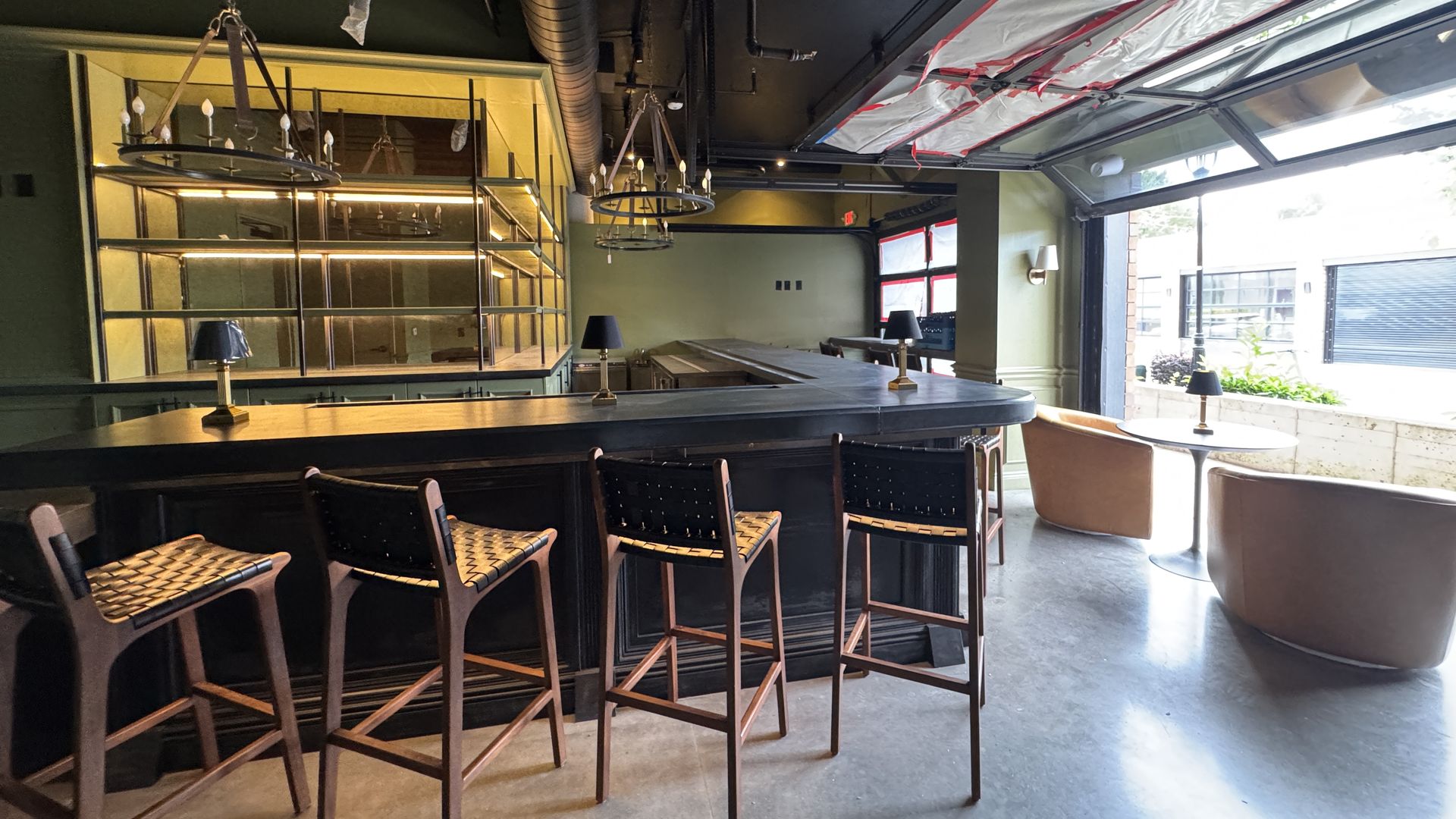 Empty bar area with dark counter, wooden high chairs, green walls, pendant lights, and large open garage-style windows letting in daylight.