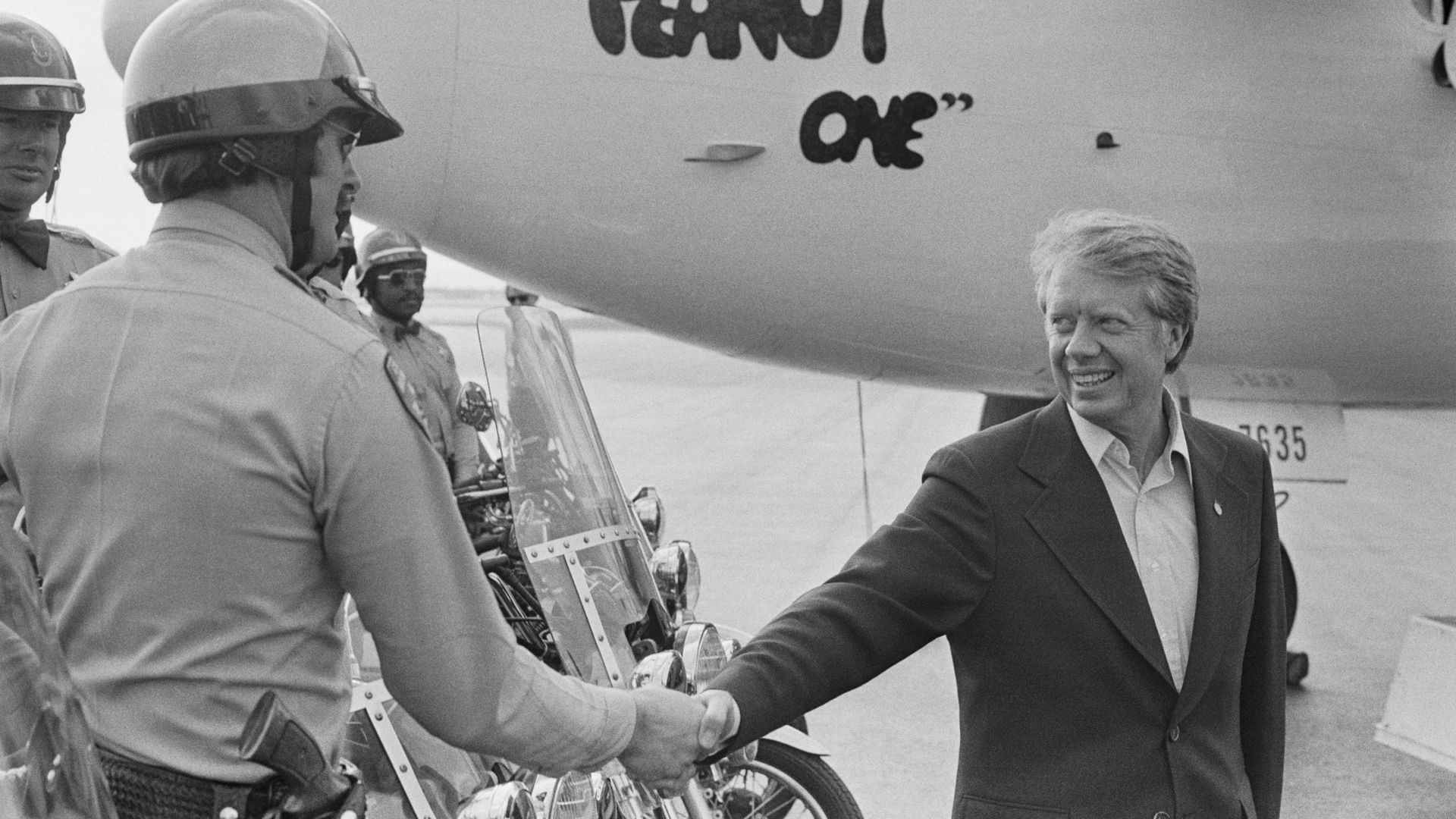 Black and white image of Jimmy Carter shaking hands with a police officer with a plane in the background with the words "Peanut One" on the nose