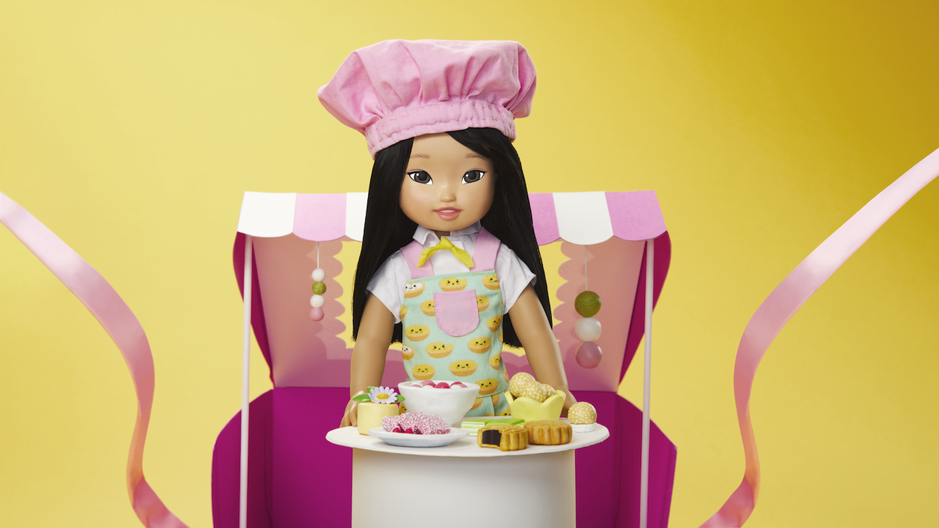 Photo of an Asian American girl doll wearing a chef's hat and holding a tray of Asian dishes