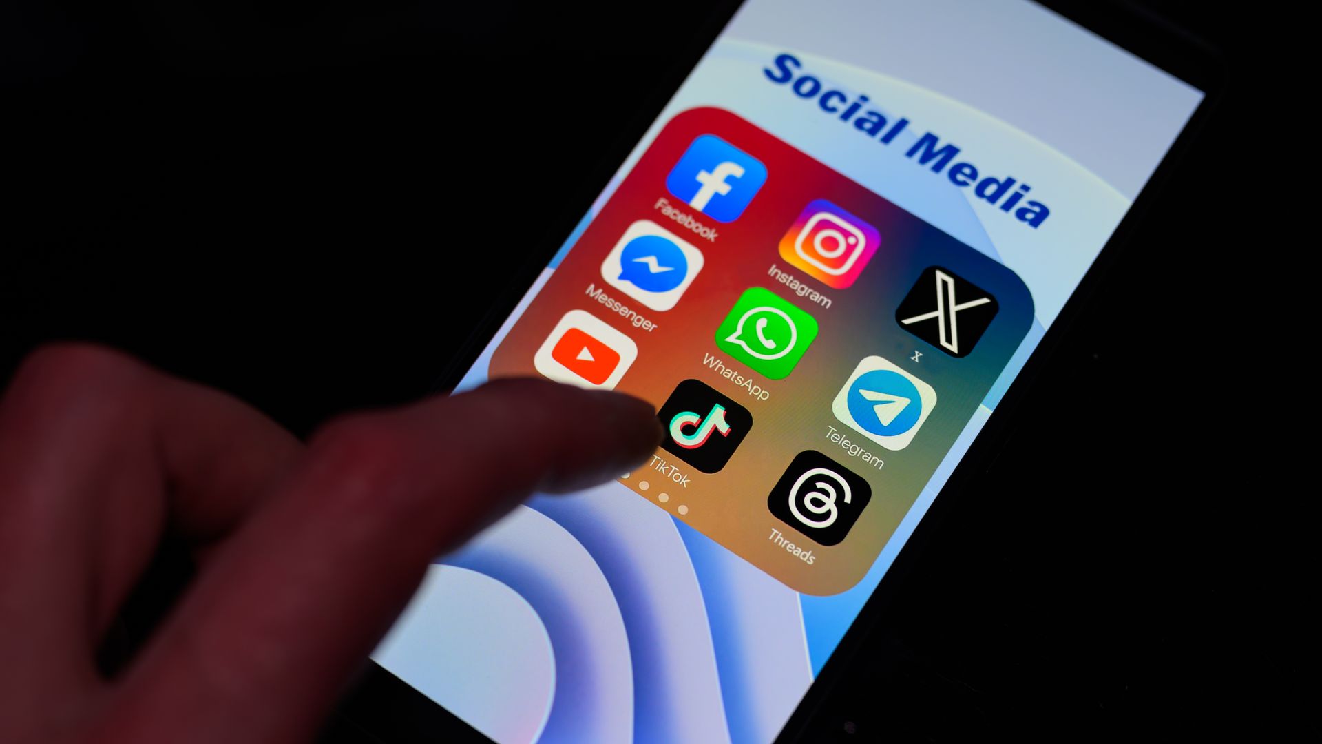 A photo of a phone screen with social media apps on the screen including Facebook, Instagram, X, TikTok and WhatsApp. A finger is starting to click on a button. 