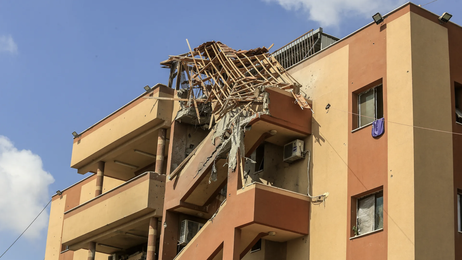 Orange and beige building with severe damage to the top corner, showing exposed wooden beams and broken concrete under a clear blue sky.