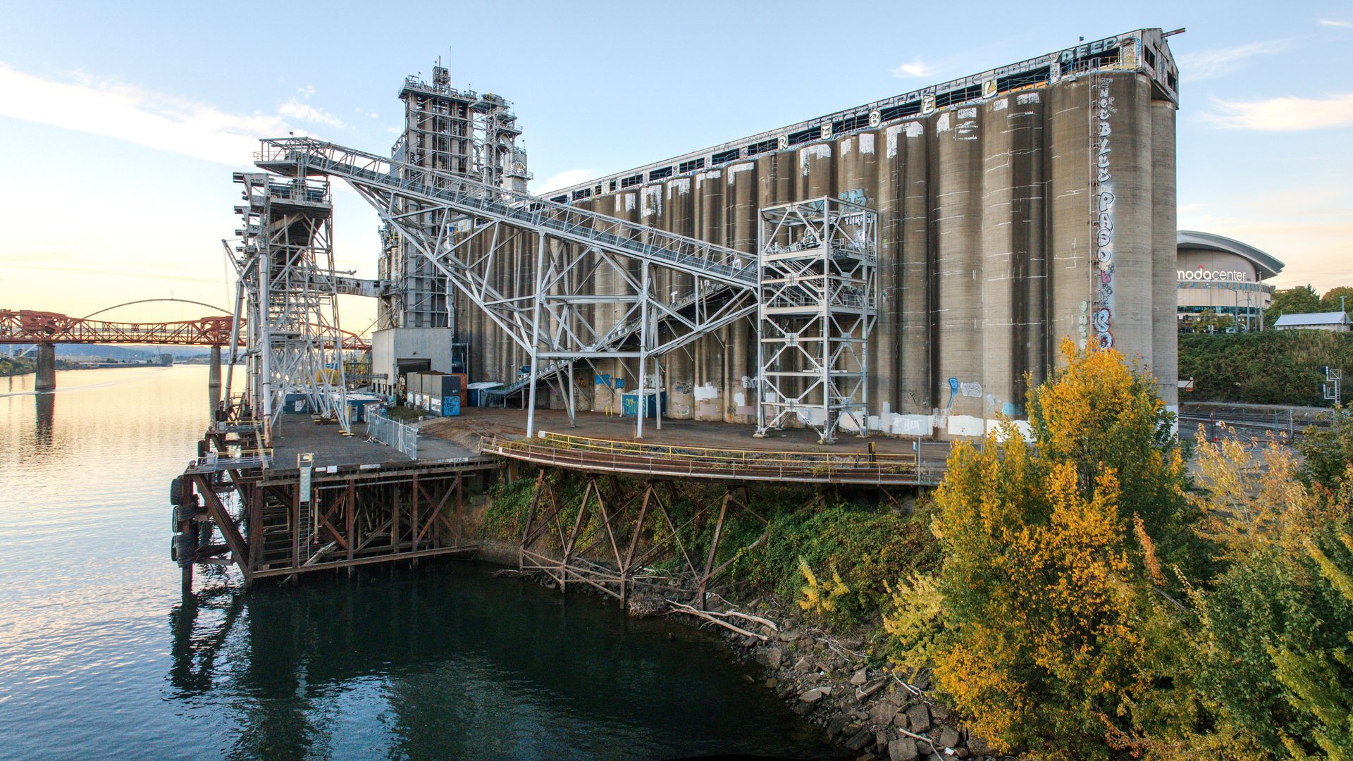 Old industrial grain silos and metal structures by the river with graffiti, autumn trees in orange and green, a red bridge over the water, and a distant curved building labeled "nodo center".