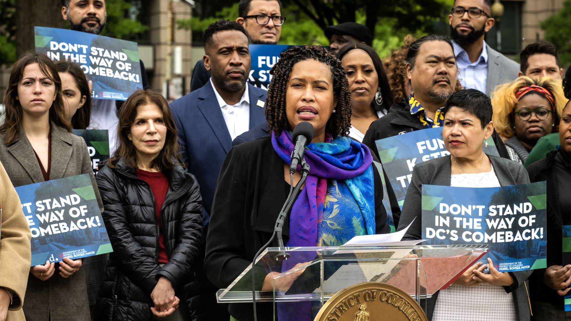 Mayor Muriel Bowser speaks from a podium in front of a crowd of people holding signs that say "Don't stand in the way of DC's comeback" at an event in Franklin Park to spark the city council to pass legislation to enliven a too quiet downtown post pandemic. 