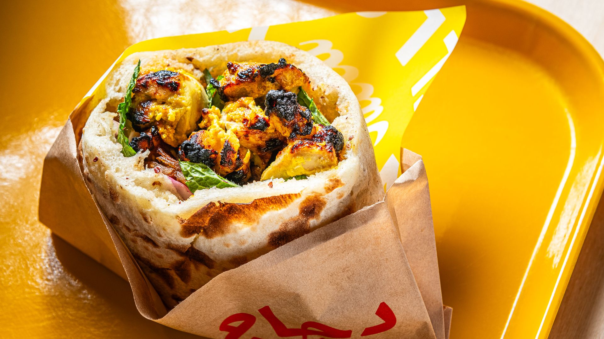 Close-up of a grilled cauliflower pita wrap with greens inside a warm flatbread, partly eaten and wrapped in brown paper, resting on a bright yellow tray.