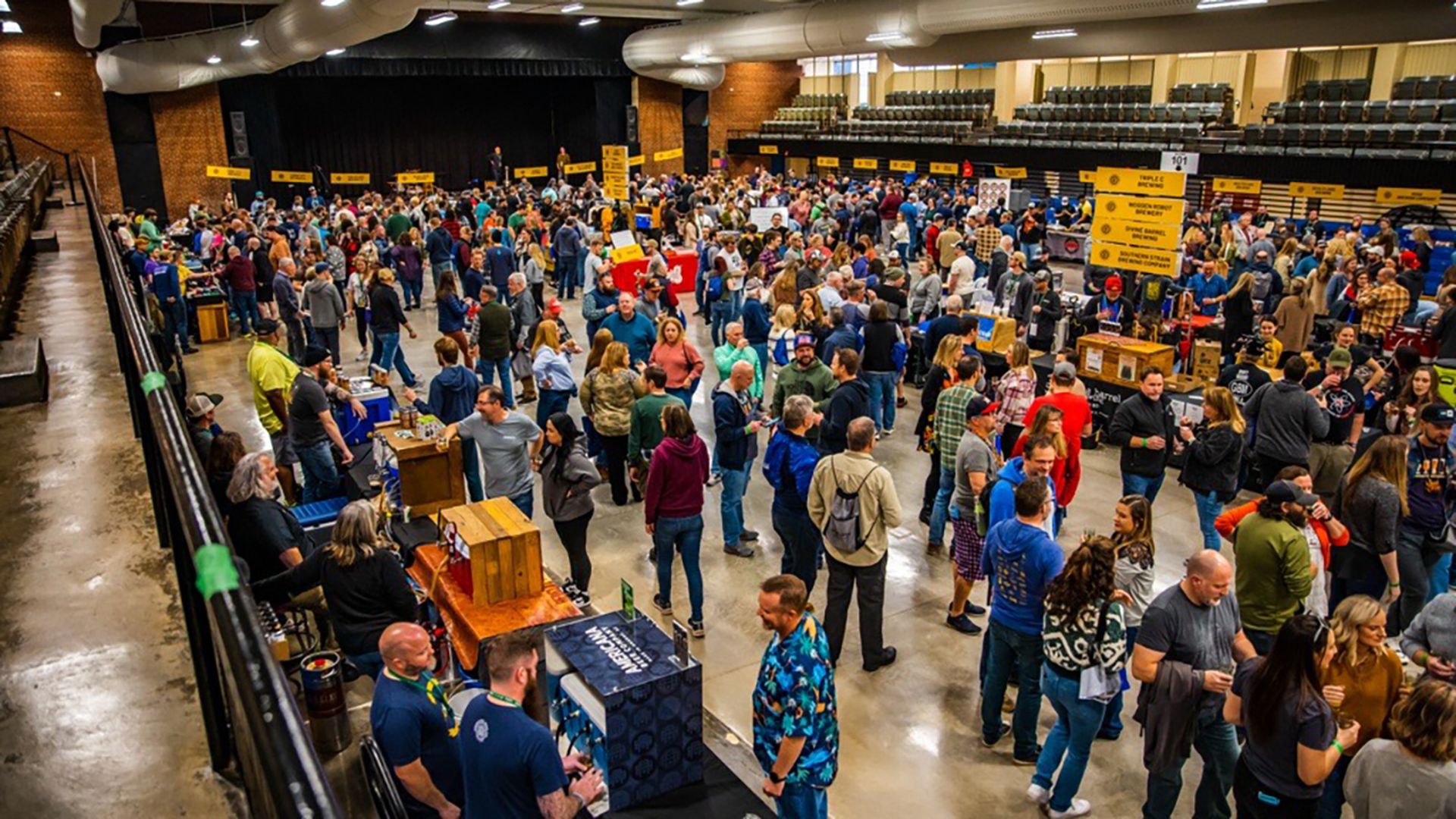 Crowded indoor event with people walking, standing, and talking around booths with yellow signs in a large hall with brick walls and rows of seats above.