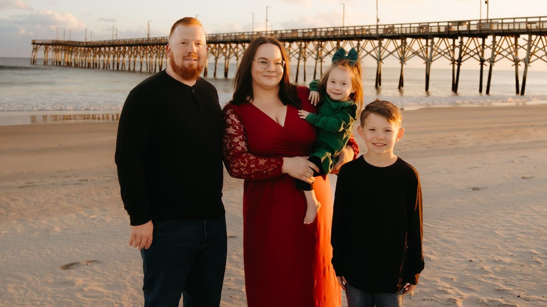 A family of four smiling together on the beach at sunset.