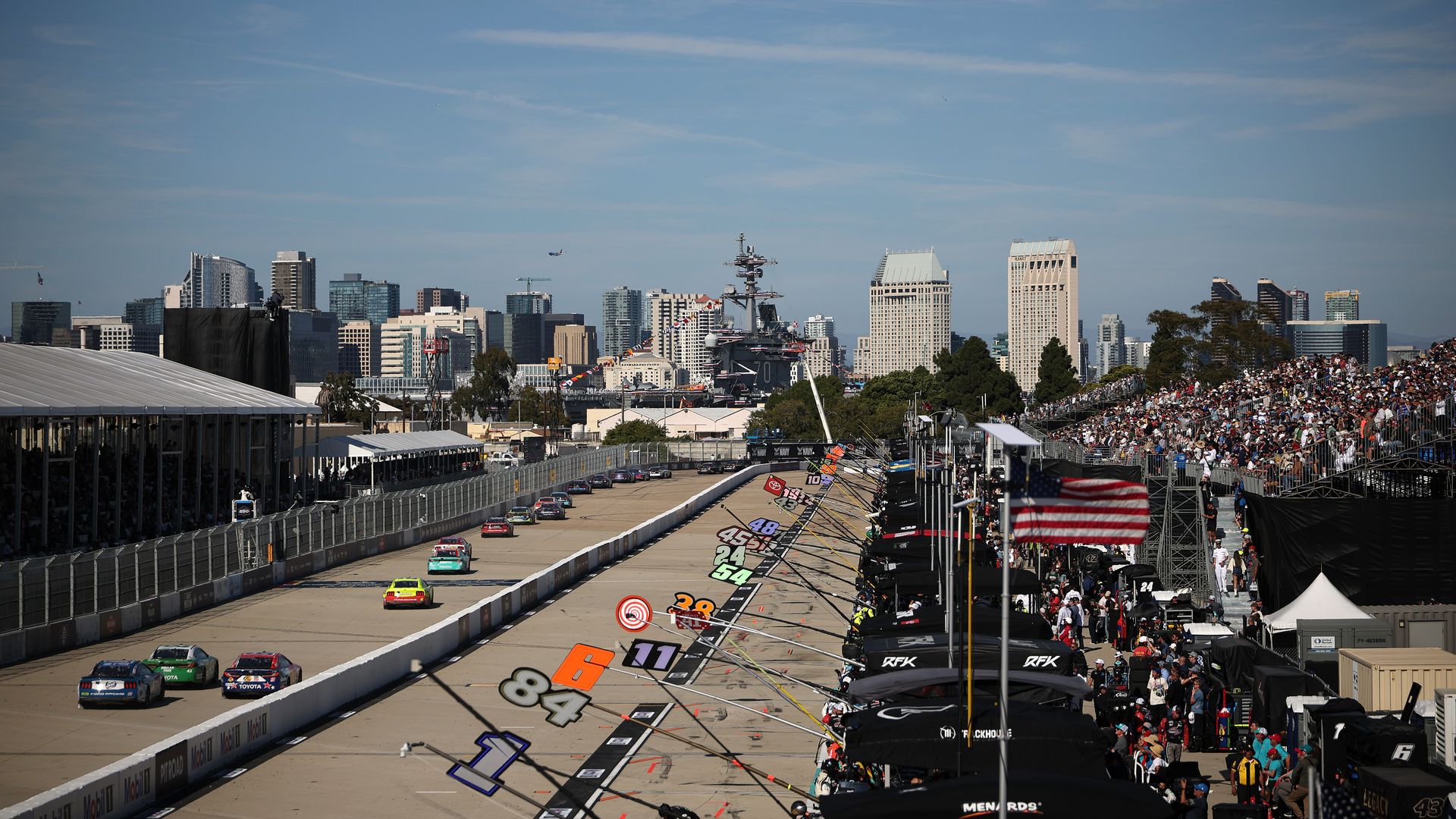 Stock cars race down a street circuit during a NASCAR event, with packed grandstands and team pit boxes lining the right side of the track. The downtown San Diego skyline rises in the background, and a large U.S. Navy aircraft carrier is visible in the harbor beyond the course under a clear sky.