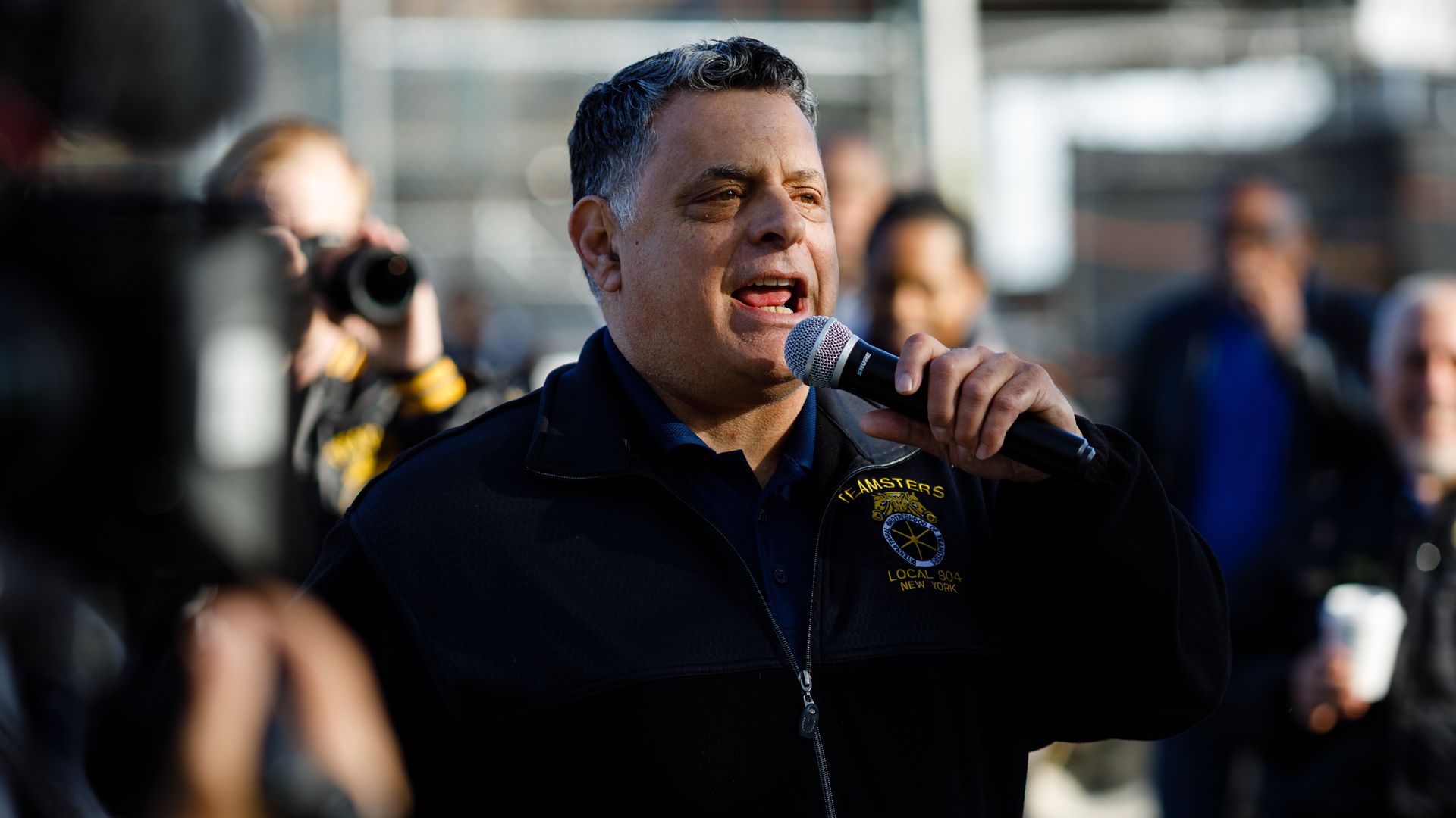 A person holding a microphone speaks outdoors