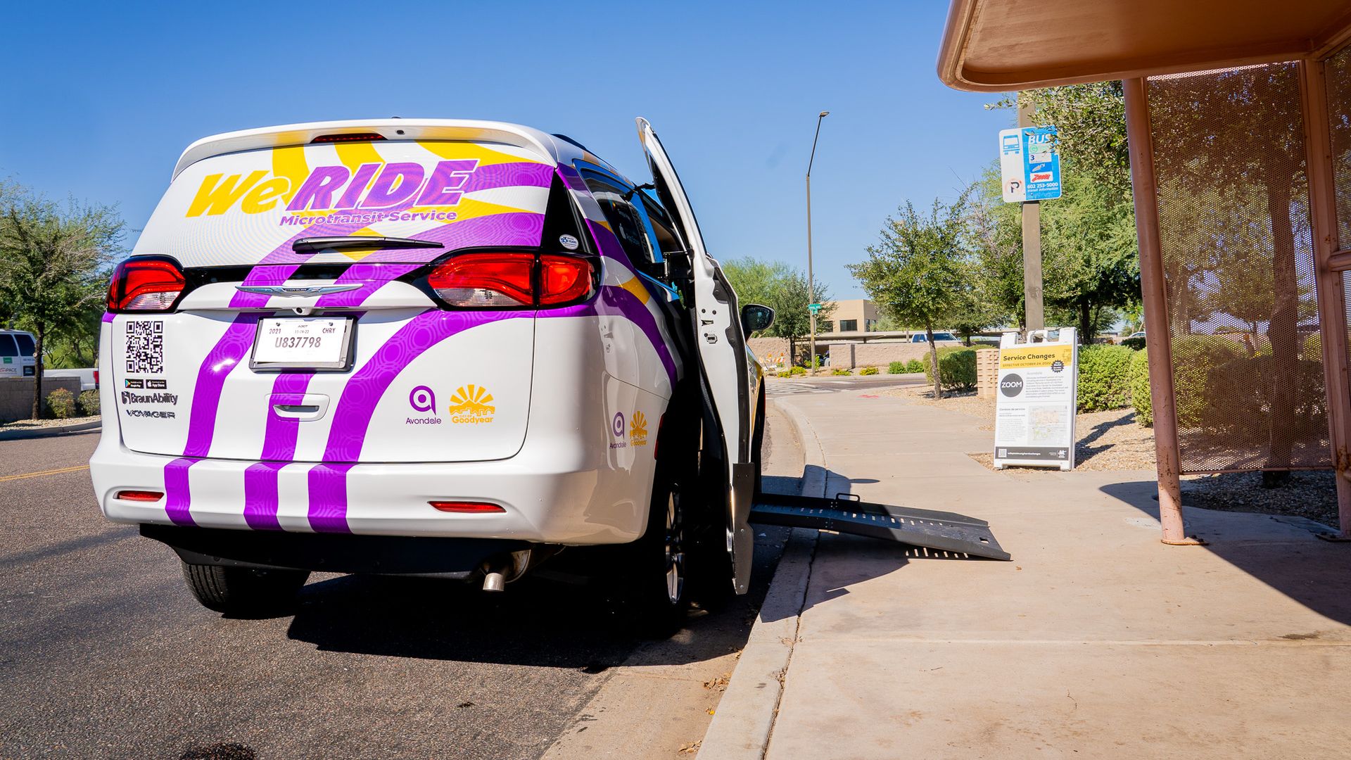 The back of a white van with purple and yellow markings with a small ramp extending from an open side door.
