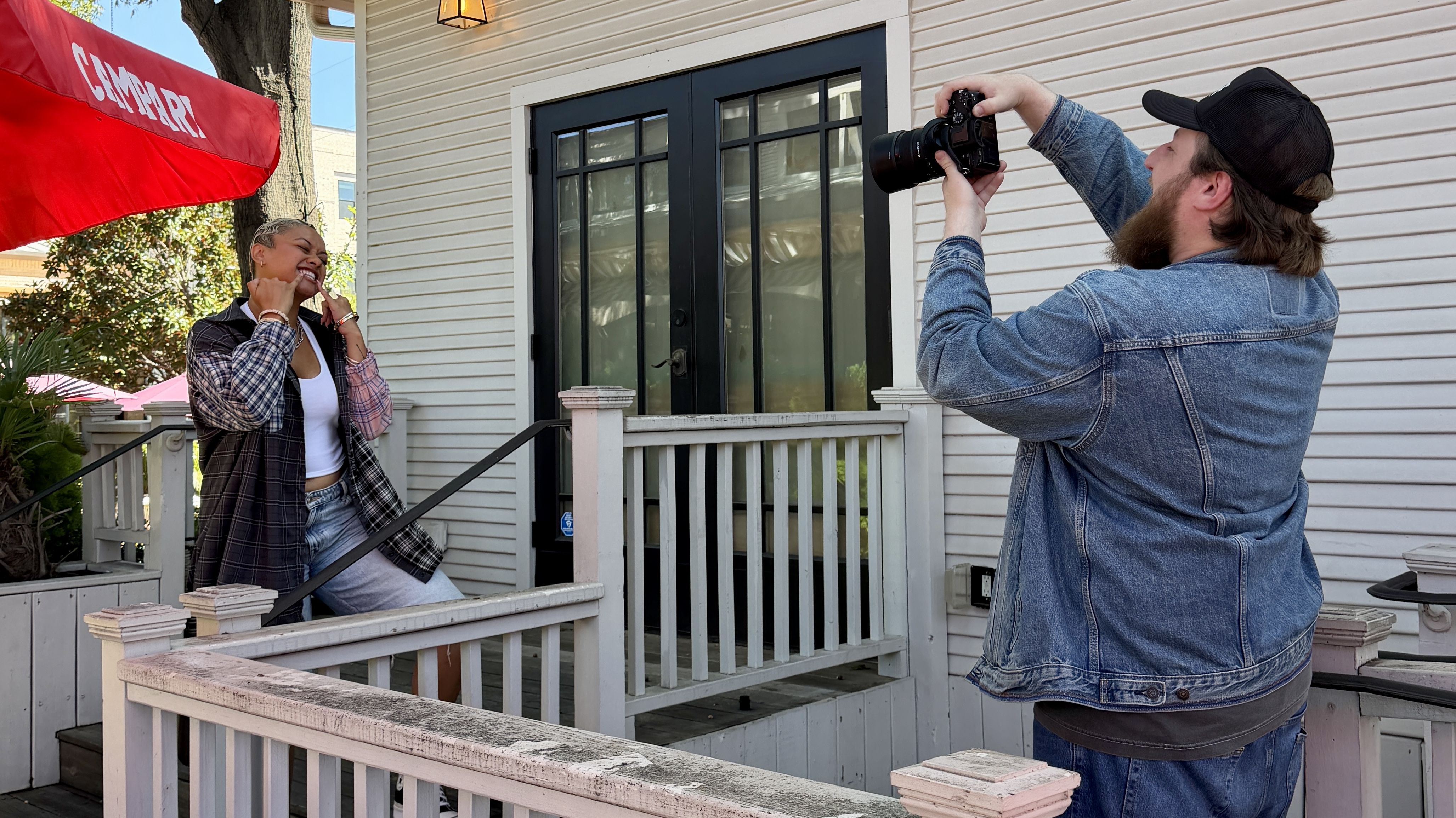 A bearded man in a denim jacket and cap films a smiling woman posing on a white porch railing, wearing a white tank top and plaid shirt, while a red canopy looms overhead.