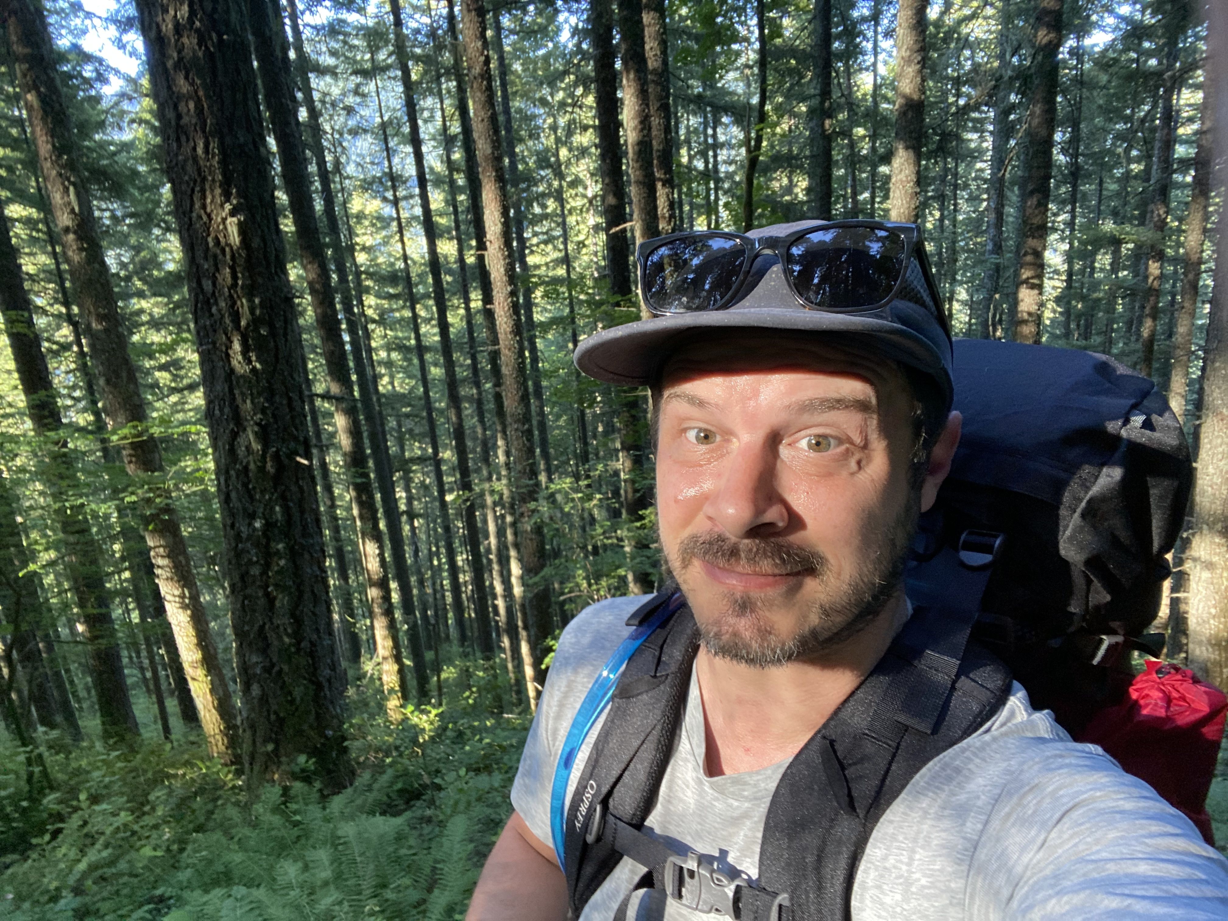 Self-portrait of a man hiking in a dense forest. He wears a gray T-shirt, a backpack, and sunglasses on his cap. Tall trees surround him as sunlight filters through the leaves.