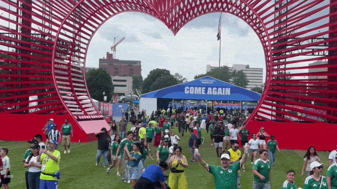 Large crowd at a park festival passes under a red heart arch; a blue tent reads COME AGAIN in the background among green shirts, people, and city buildings.
