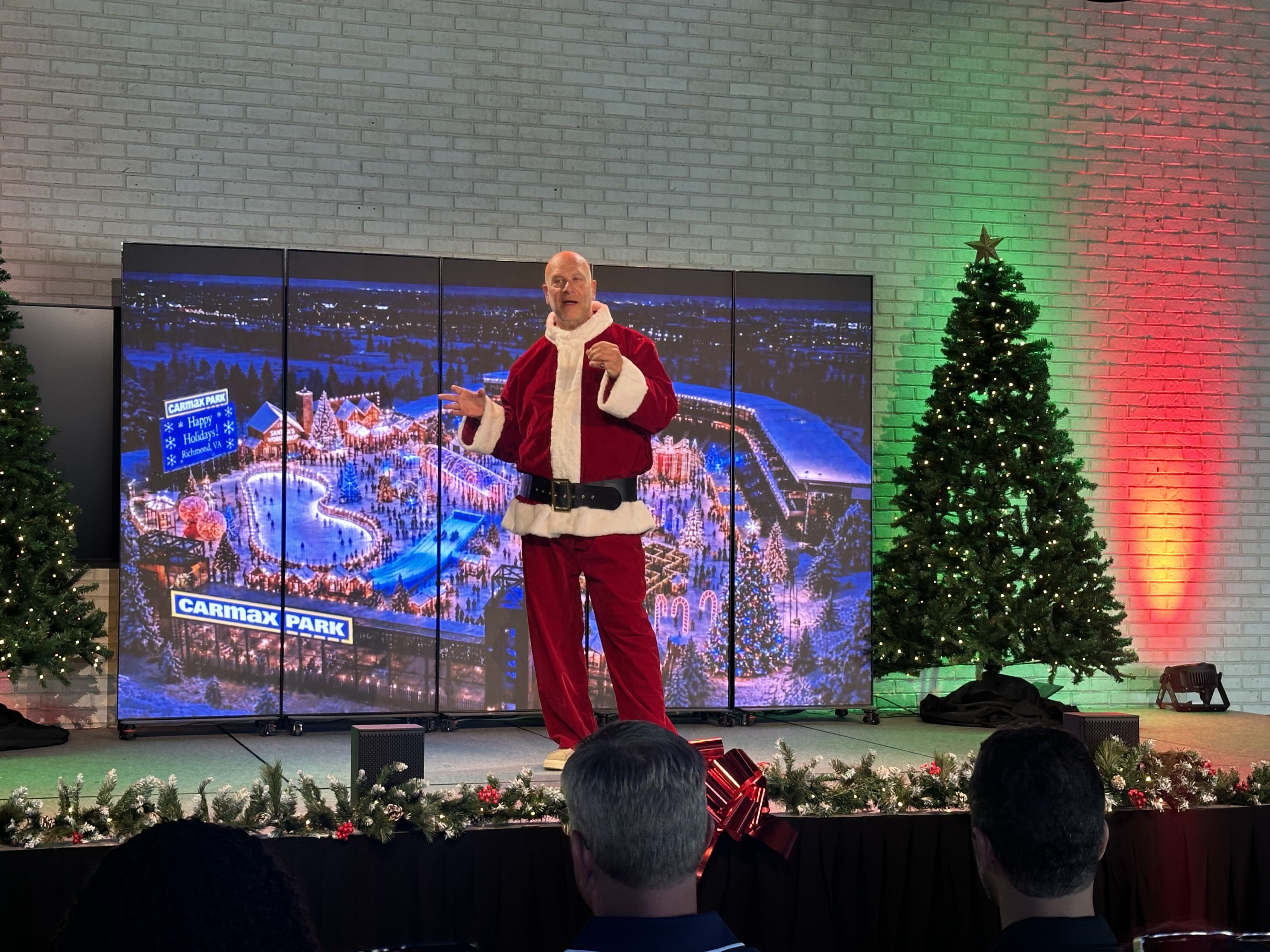 Santa in a red suit stands on a stage before a multi-panel screen showing a snowy Christmas park labeled "Carmax Park". Green and red lights illuminate brick walls as Christmas trees glow; audience watches.