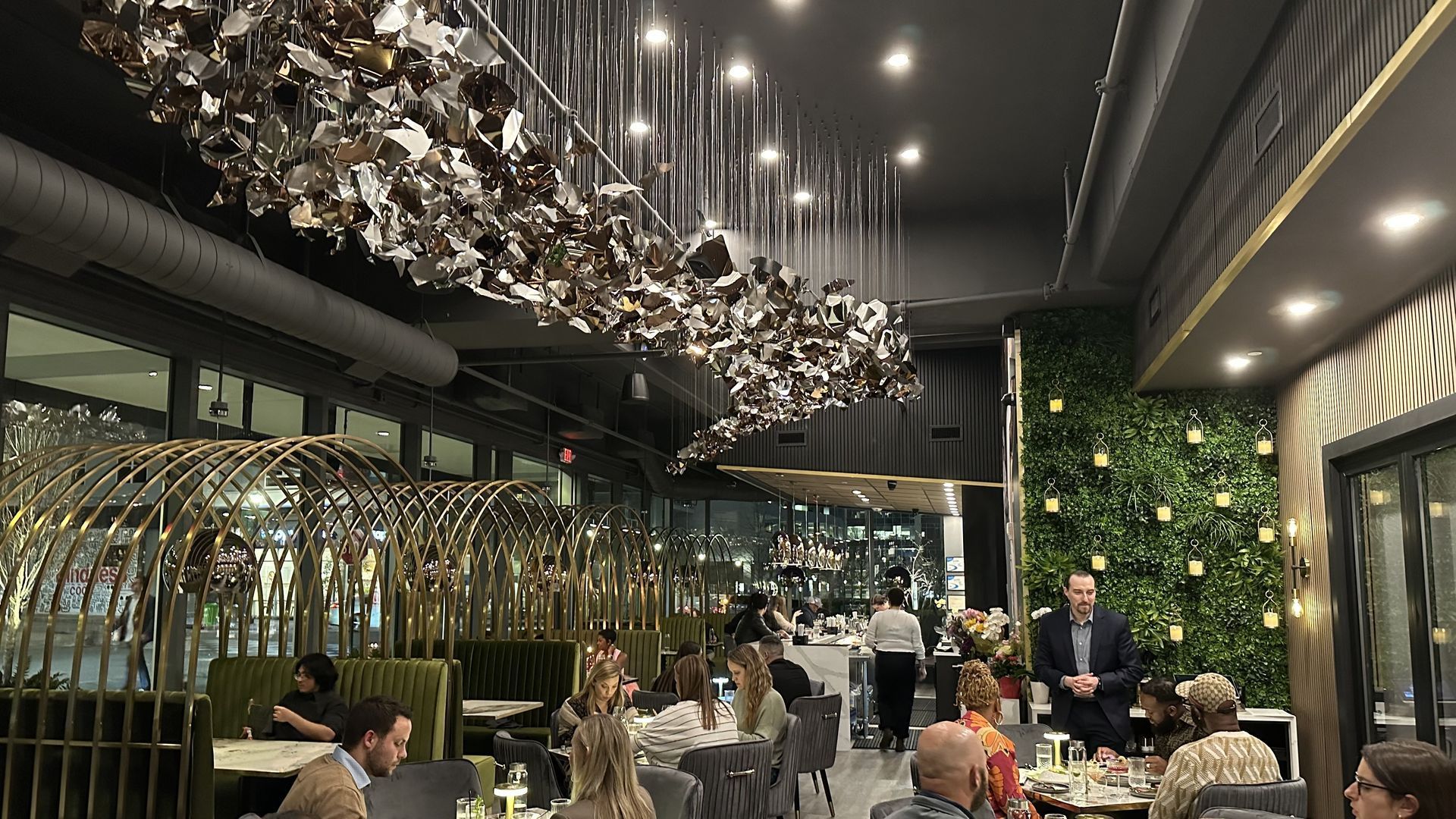 Modern restaurant interior with a ceiling tapestry of metallic origami shapes hanging from cords, lit by string lights; a green living wall with lanterns on the right and diners at tables.