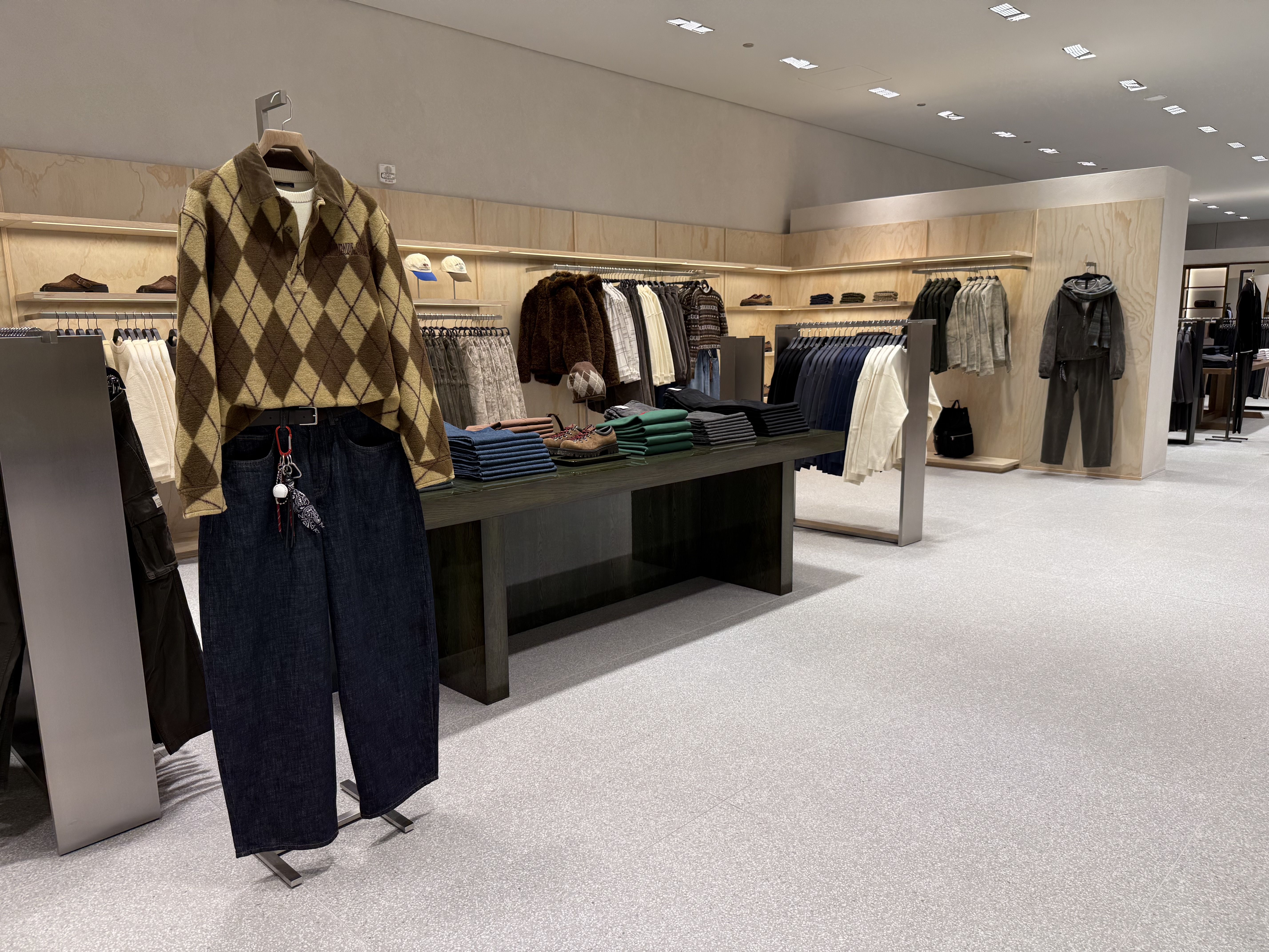Clothing store interior with beige and brown argyle sweater paired with dark wide-leg jeans on a mannequin. Shelves and racks display folded clothes and jackets in neutral tones.