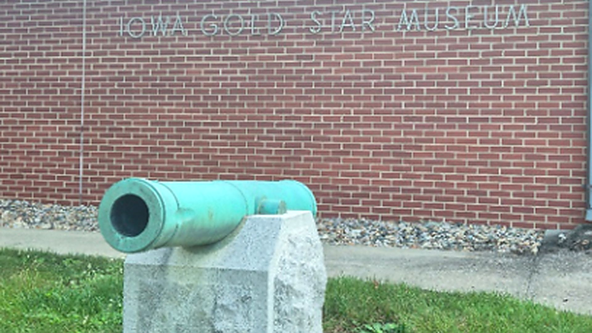 Turquoise cannon on a gray stone pedestal sits on grass, facing left. In the background is a red brick wall with faded lettering "IOWA GOLD STAR MUSEUM".