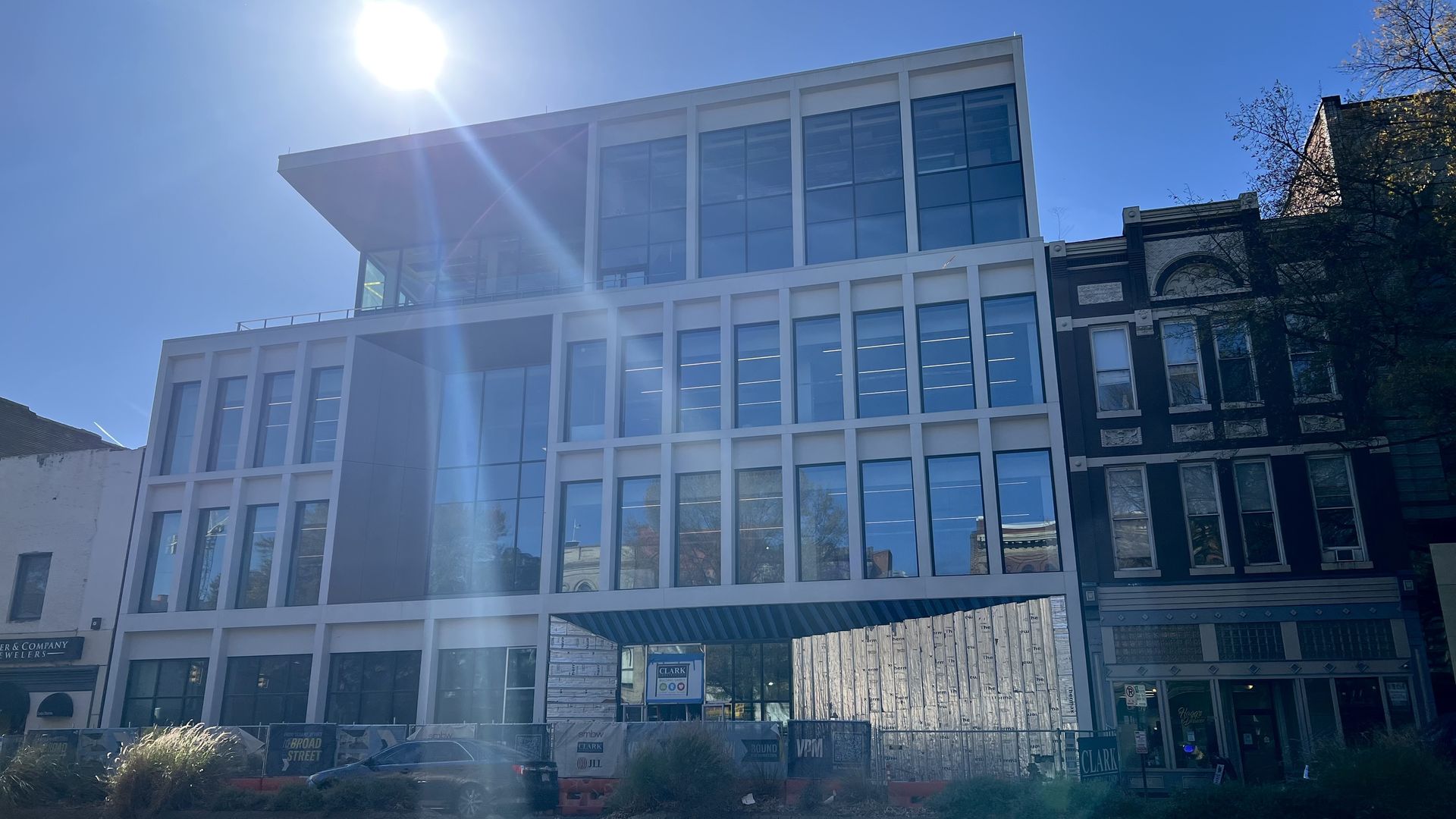 Modern white building with large blue-tinted windows, under bright sunlight and clear blue sky, with construction fencing and neighboring older buildings on the sides.