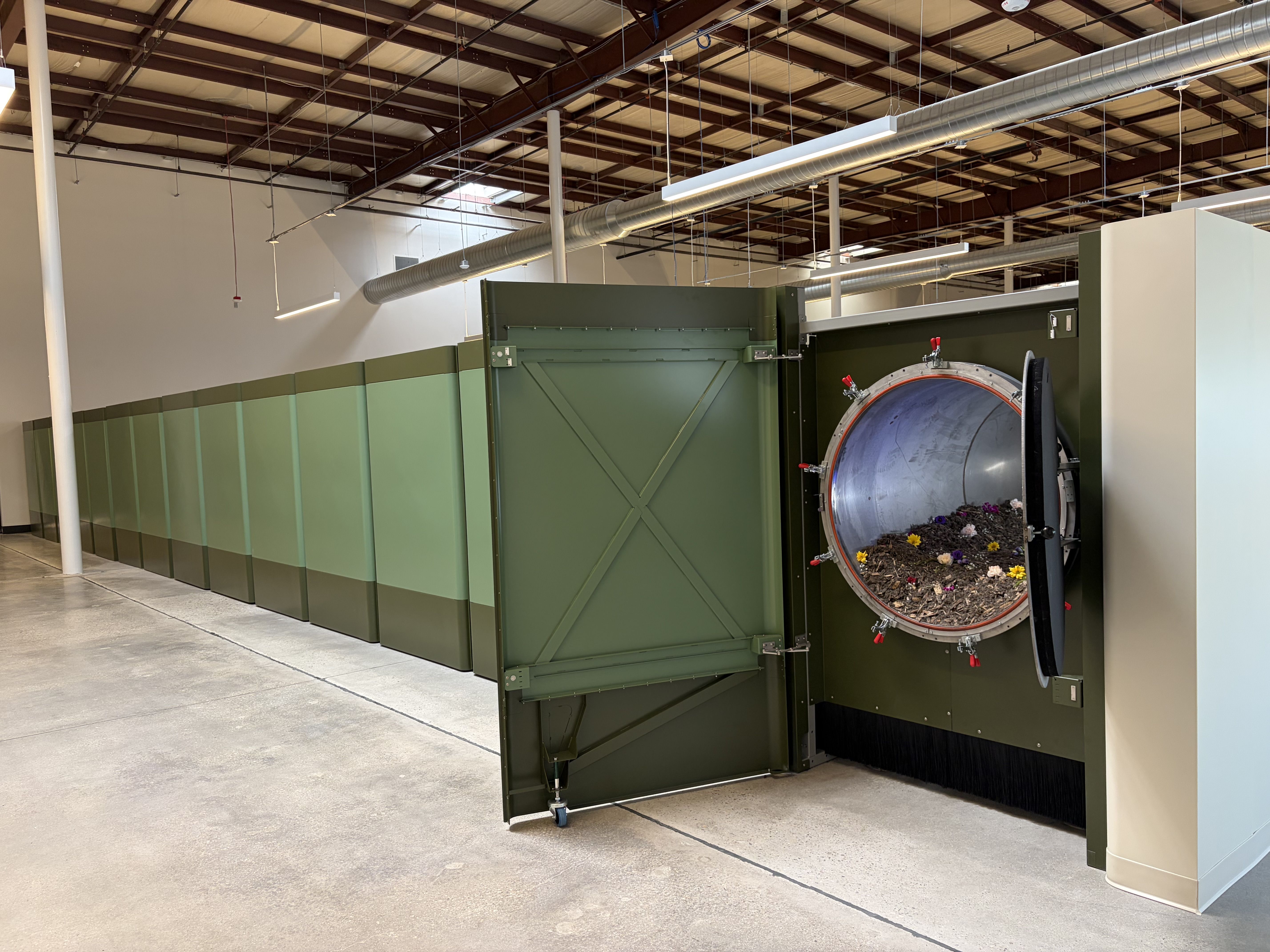 Industrial interior with exposed brown steel beams and hanging ducts. A large green machine with a circular open door shows soil and colorful flowers inside; pale green wall panels line the room.