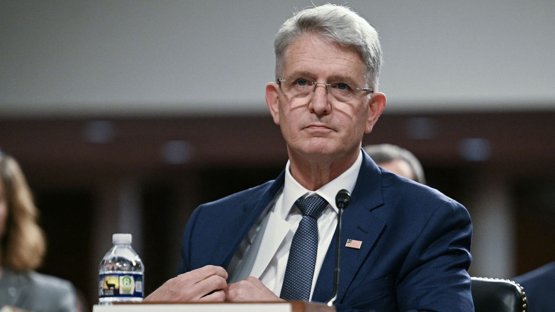 A man in a blue suit, white shirt and blue tie sits behind a desk during a congressional hearing. A water bottle is seen on the table to his right.