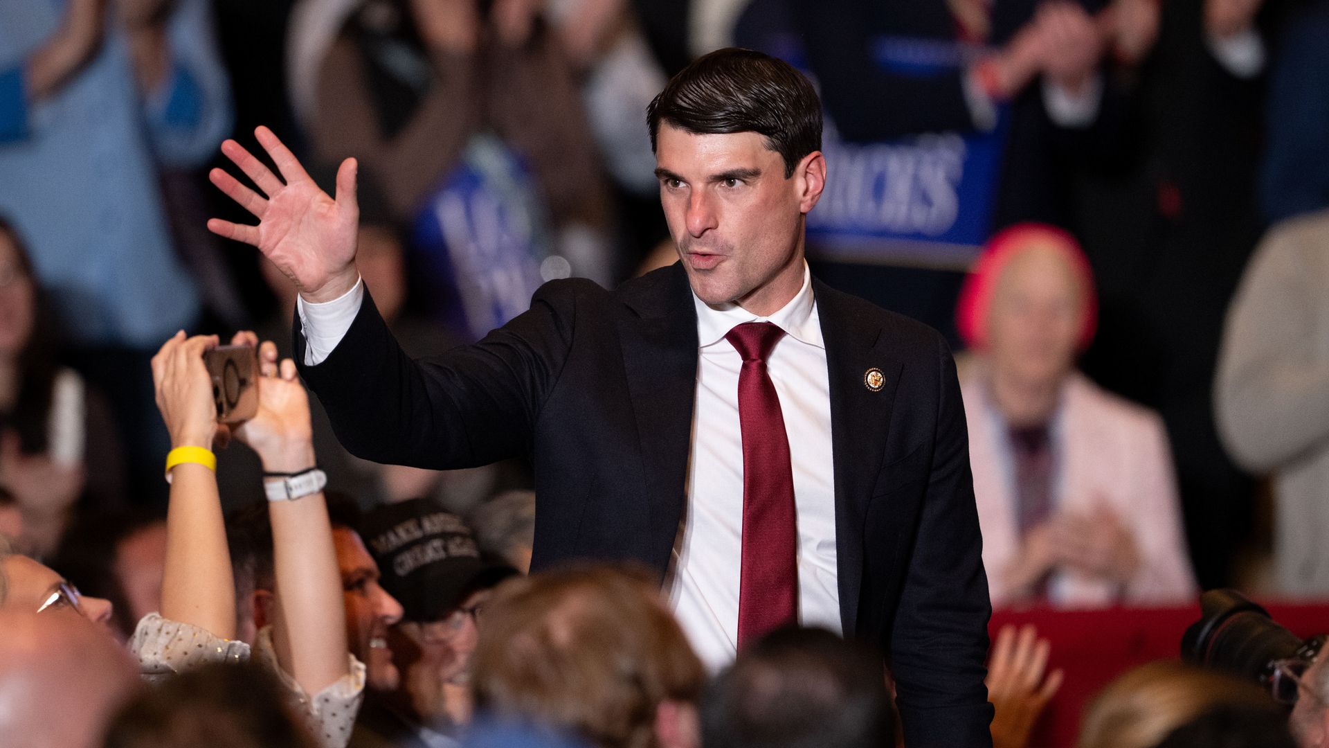 A man in a dark suit and red tie raises his hand to greet a cheering crowd at a political event, with fans, photographers, and raised hands surrounding him.
