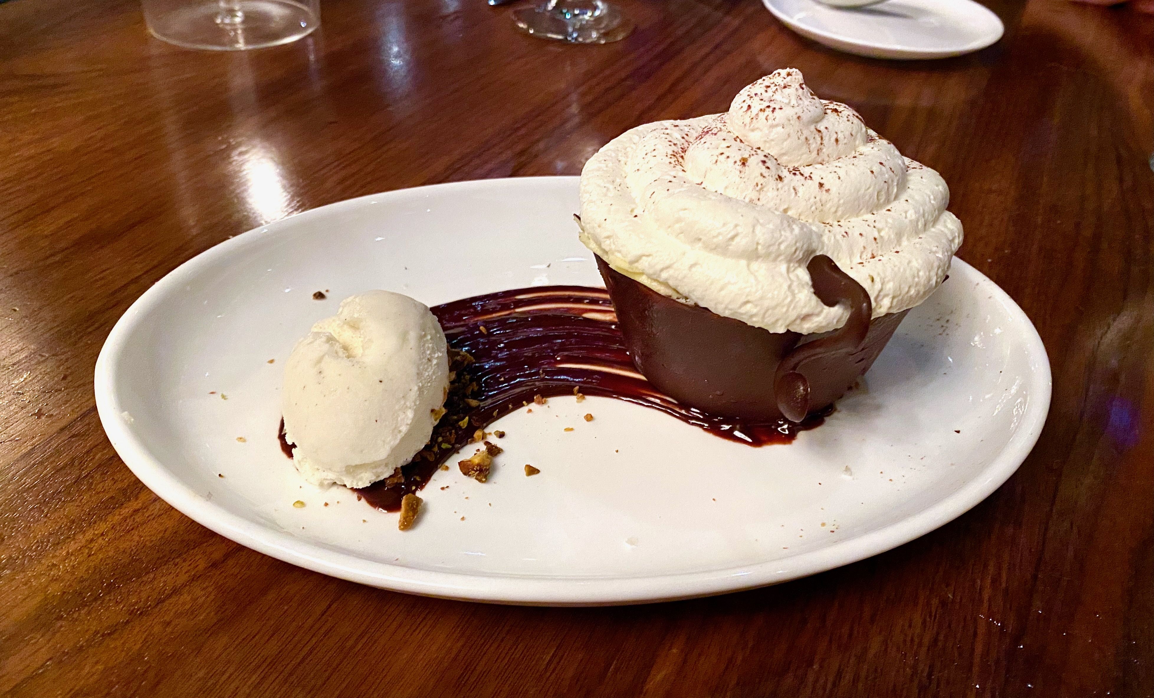 Tiramisu on a white plate inside a chocolate teacup, with whipped cream on top