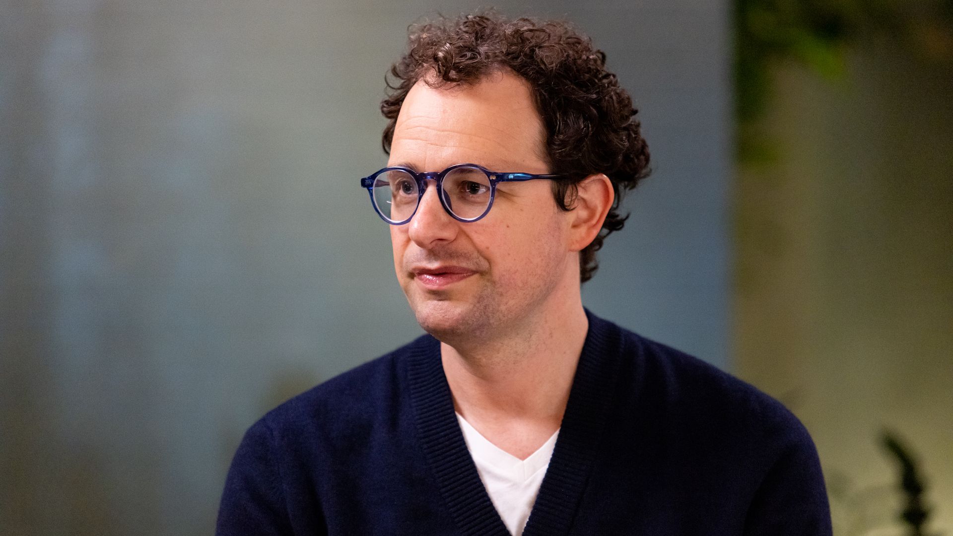 A man in glasses and a cardigan sitting down and staring to the left. 