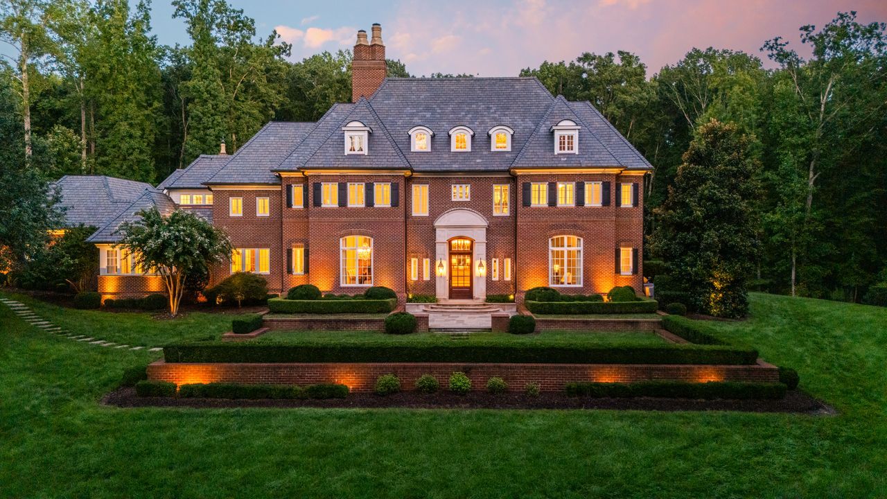 Large brick mansion with lit windows at dusk, surrounded by green lawn and trees, featuring a gray slate roof and landscaped shrubs with warm exterior lighting.