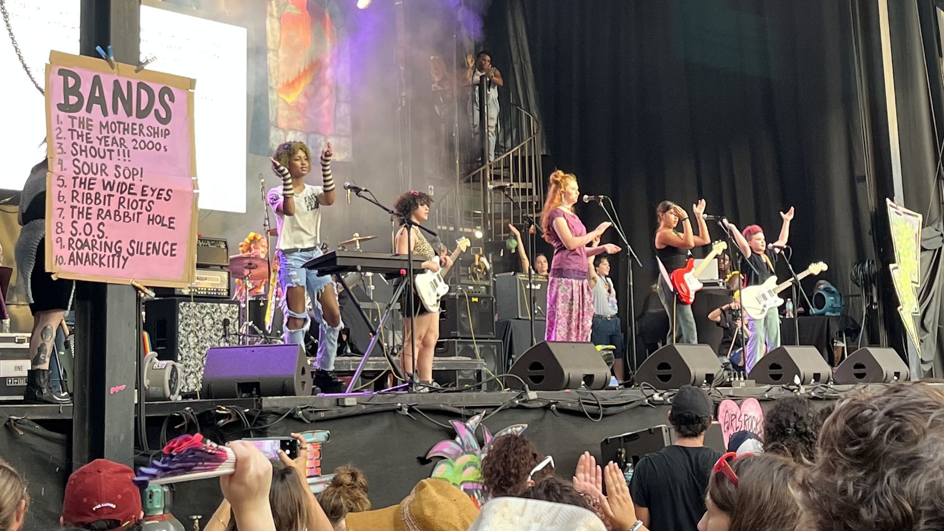 Outdoor concert scene with a diverse band performing on stage; a large pink sign labeled 'BANDS' lists songs; purple lighting, guitars and microphones, and a cheering crowd wearing hats.