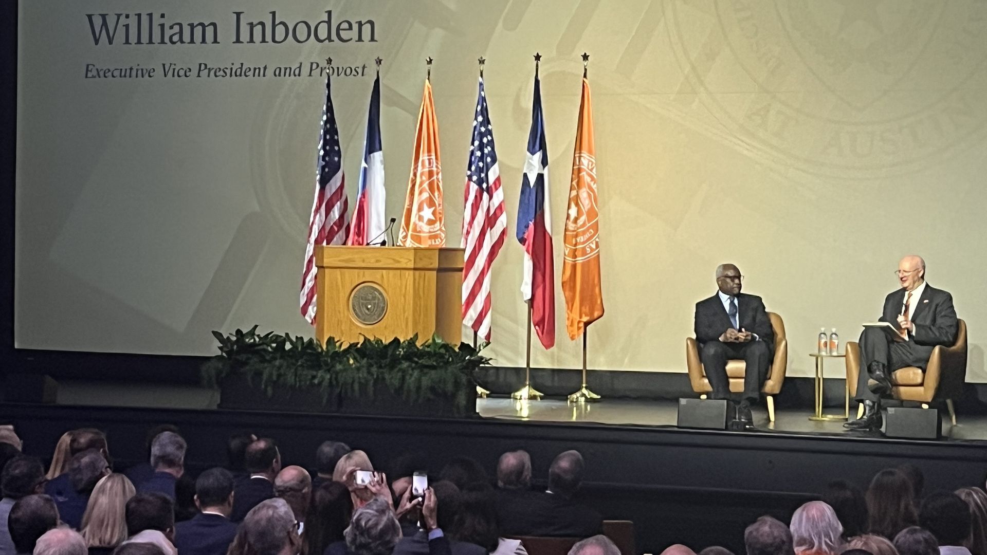 Stage with a wooden podium, US and other flags behind it; two men in suits sit in chairs on the right, audience in foreground; large backdrop shows names and titles.