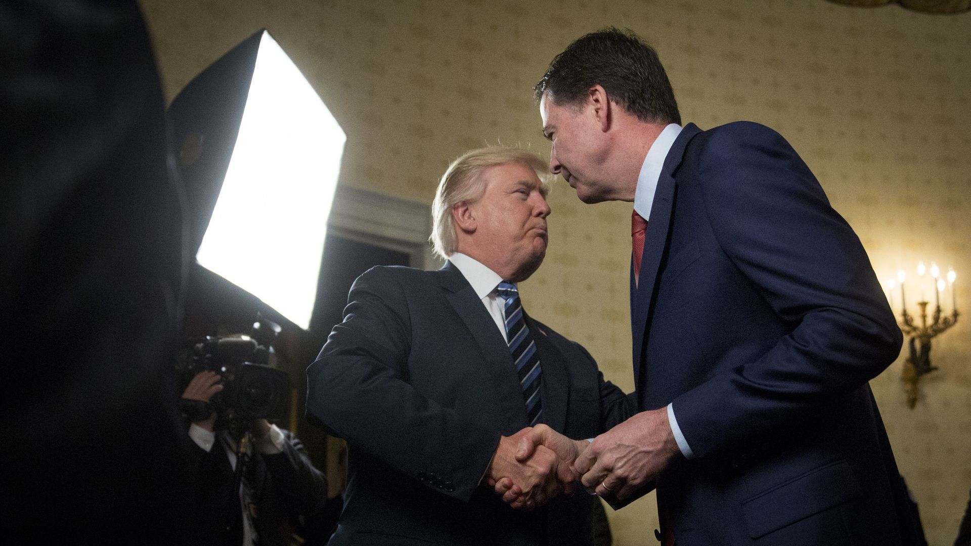 Trump shakes hands with James Comey. Both men are wearing suits and a large light shines on them as a person holding a camera captures the moment.